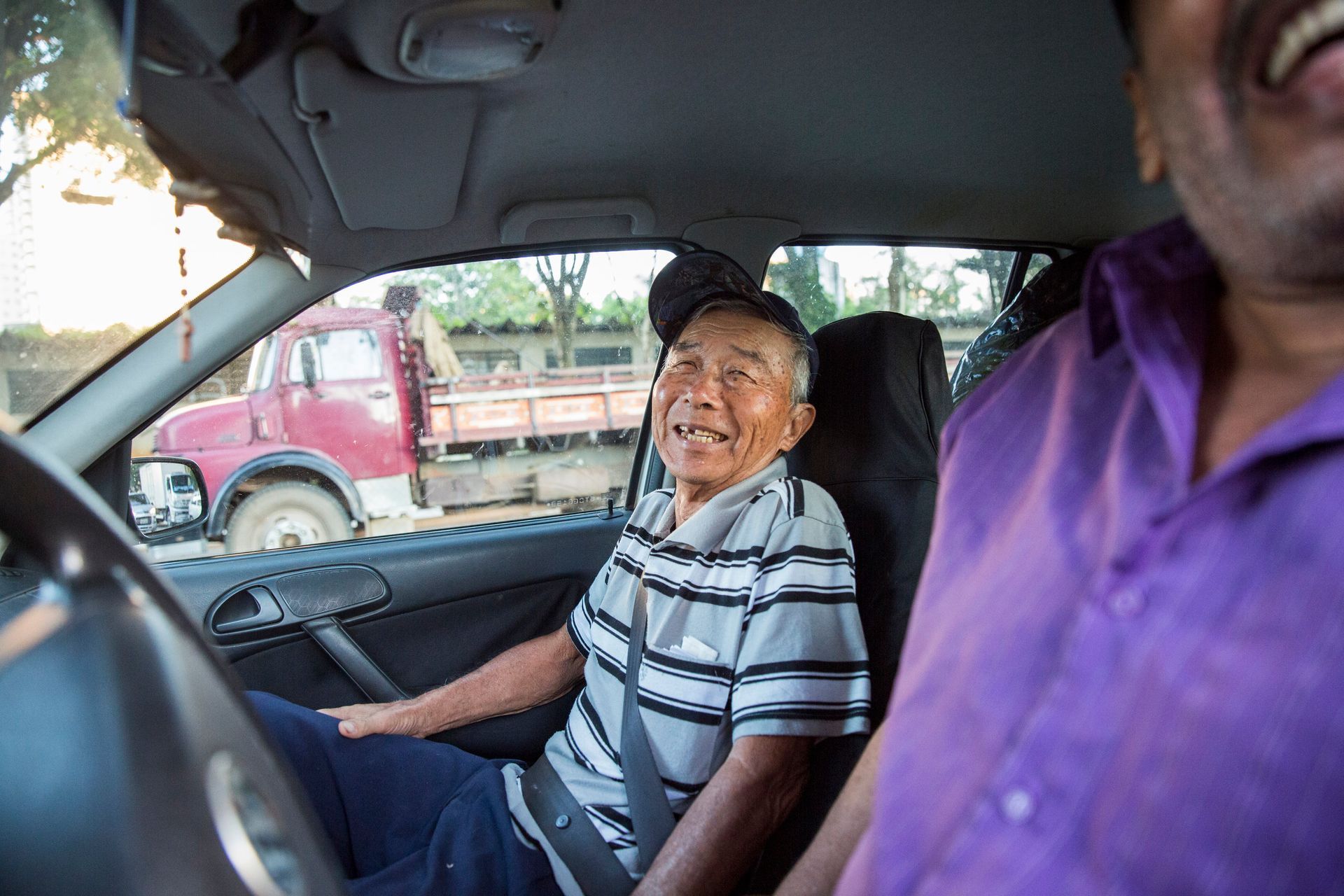 Um homem sorridente de camisa listrada está sentado em um carro, com uma pessoa de camisa roxa em primeiro plano.