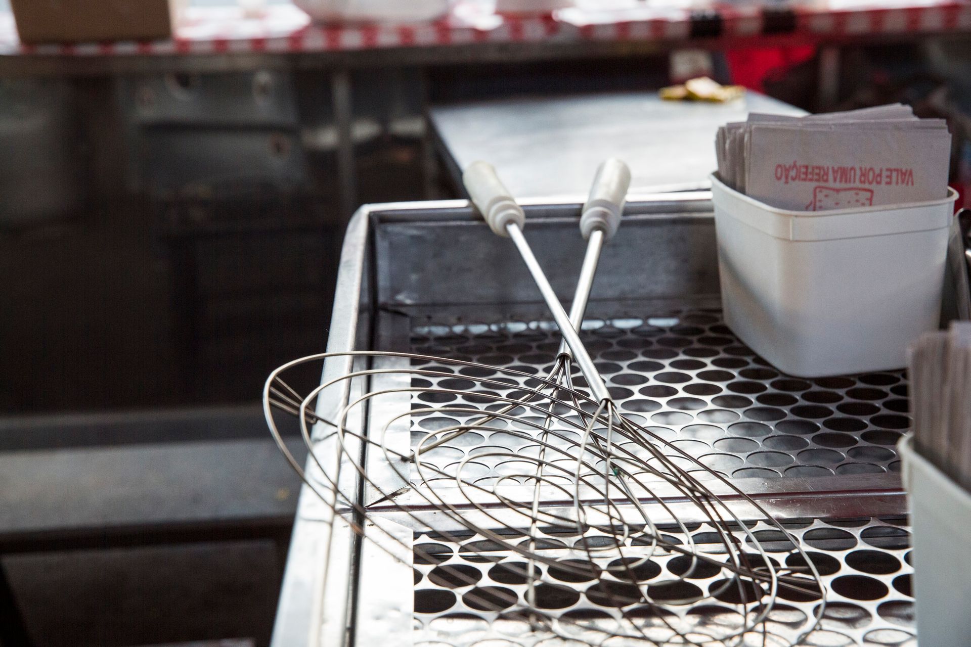 Um escorredor de arame e utensílios sobre a bancada da cozinha, ao lado de um recipiente branco com inscrições em vermelho.