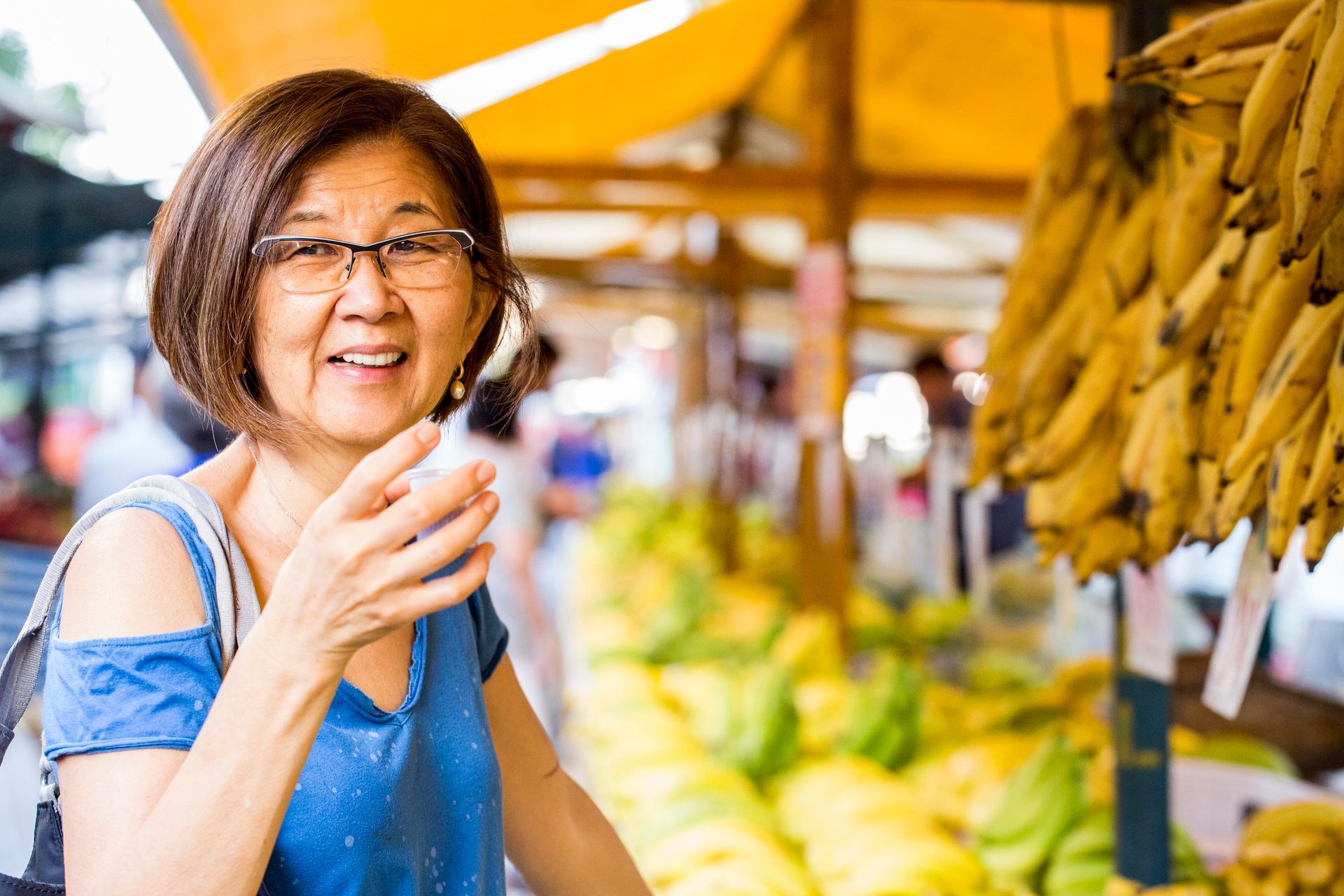 Compradora sorridente segurando uma sacola em um mercado colorido ao ar livre, com bananas penduradas ao lado.