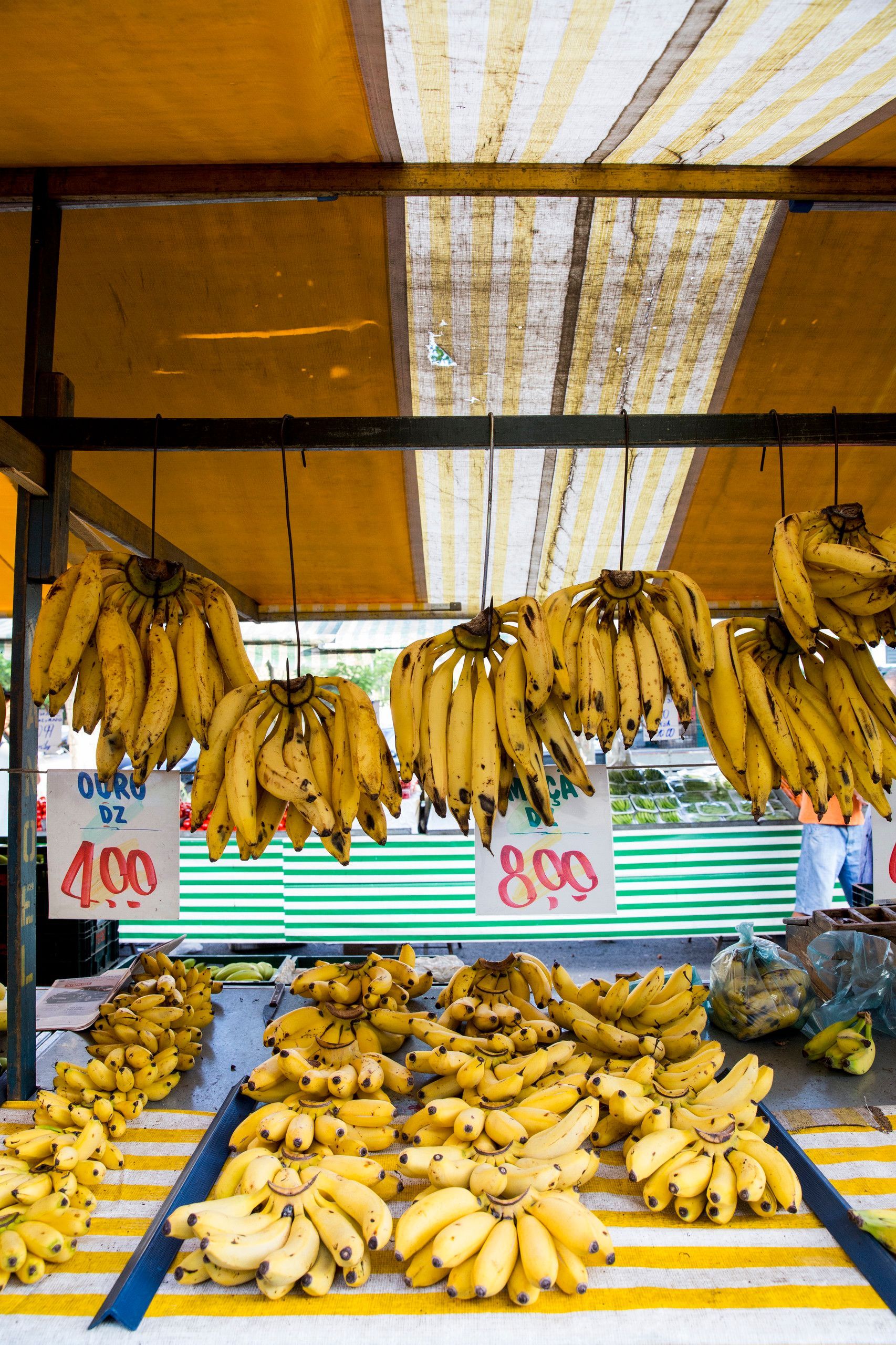 Bananas empilhadas e penduradas em uma barraca de mercado sob um toldo amarelo, com placas de preço visíveis.