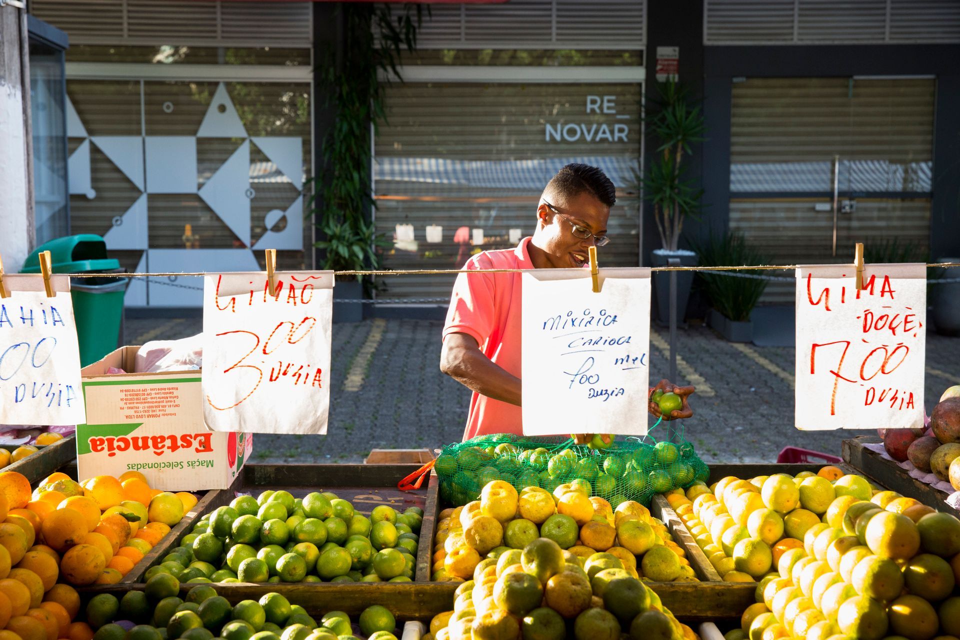 Vendedor atrás de uma banca de frutas colorida com cartazes escritos à mão na frente de uma loja.