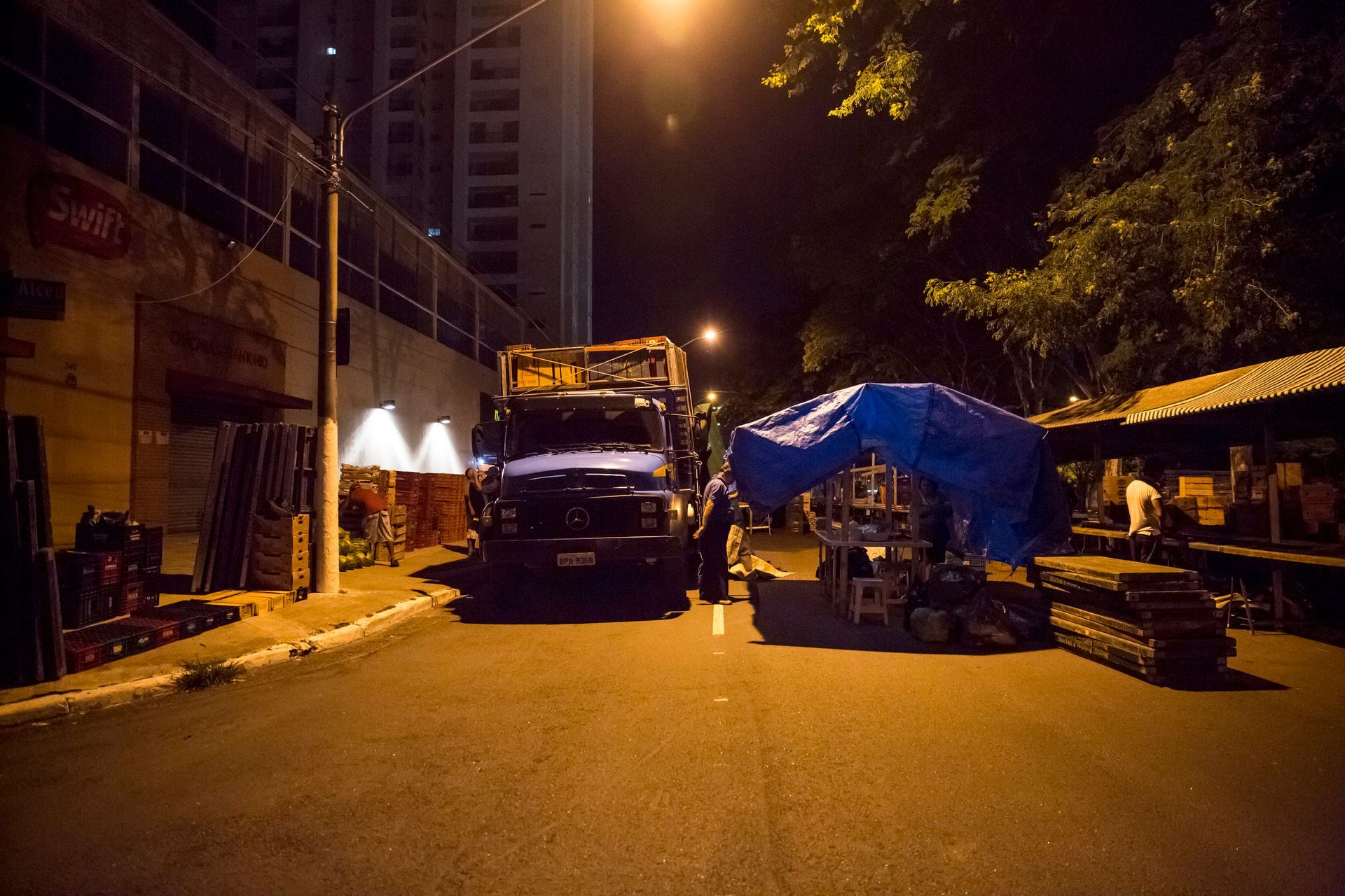 Rua à noite com um caminhão estacionado sob postes de luz e barracas de mercado cobertas por lonas azuis.