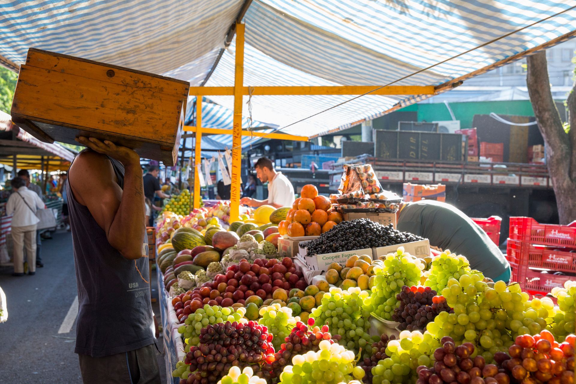 Banca de mercado a céu aberto com pilhas de frutas coloridas e um vendedor carregando uma caixa no ombro.