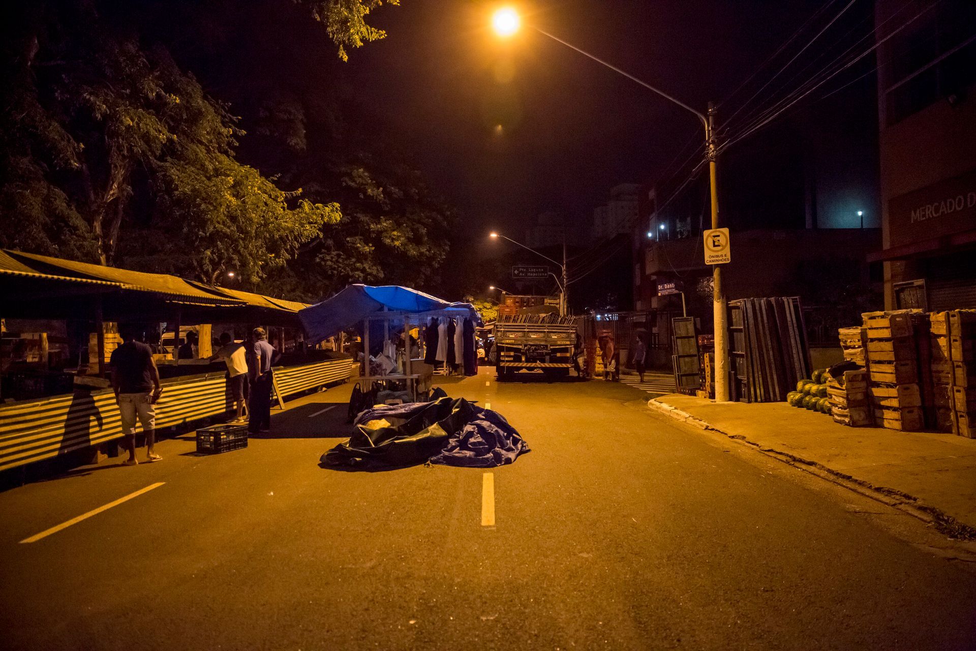 Cena noturna de rua com barricadas e lonas bloqueando uma estrada mal iluminada por postes de luz alaranjados.