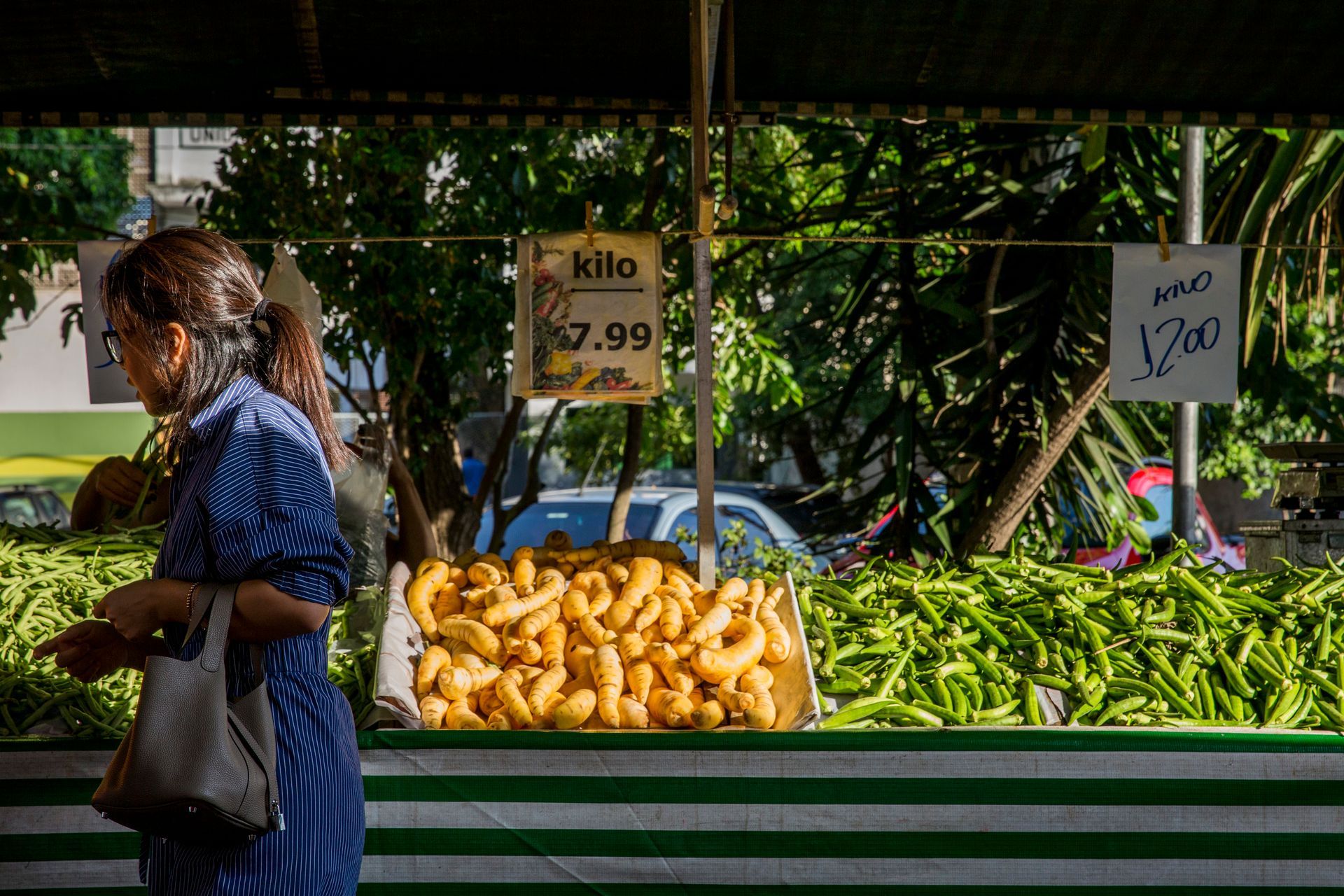 Banca de mercado com bananas, pimentões verdes e placas de preços; um comprador passa em primeiro plano.