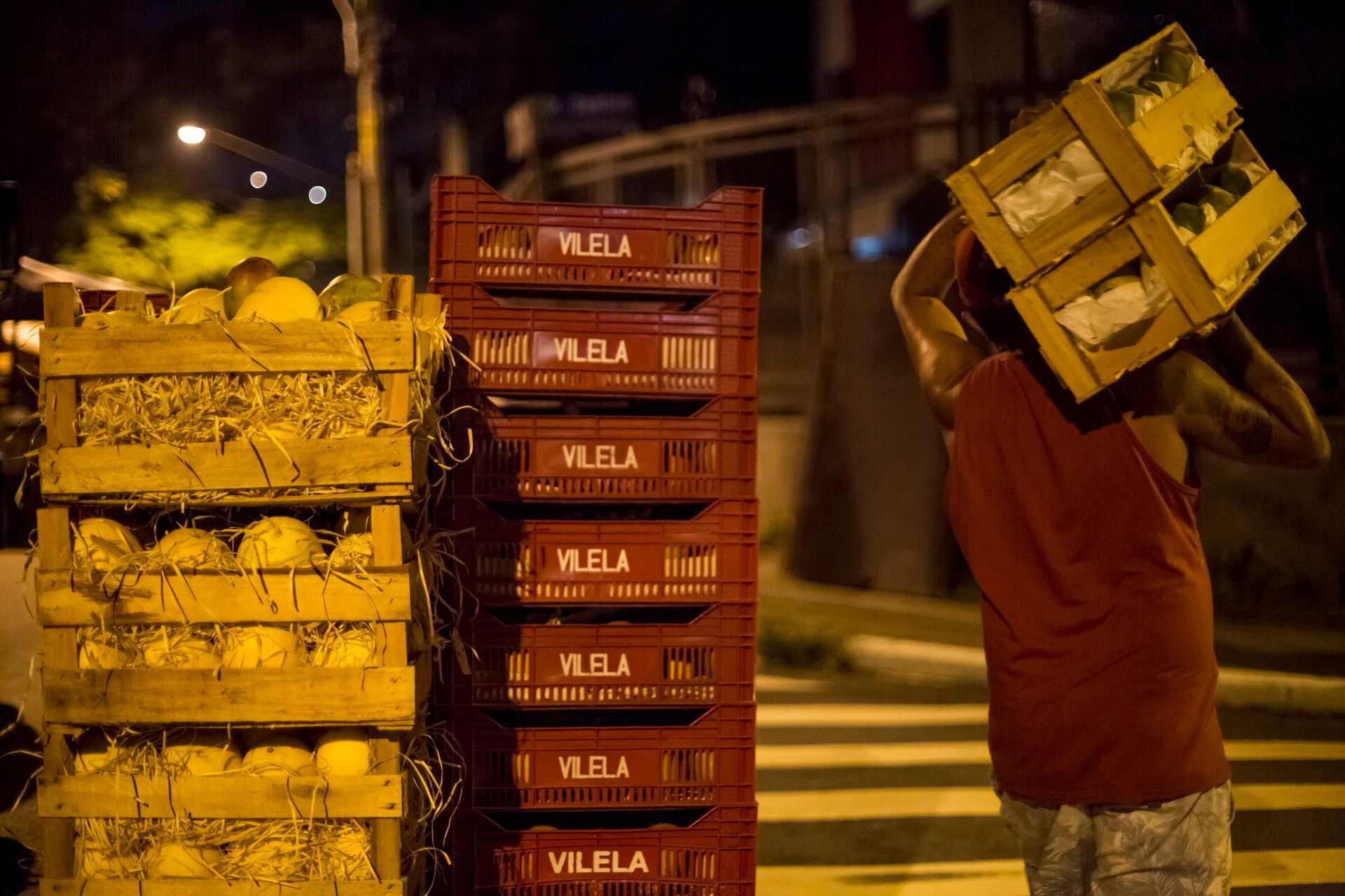 Trabalhador de mercado noturno carregando caixas de produtos em uma rua pouco iluminada.