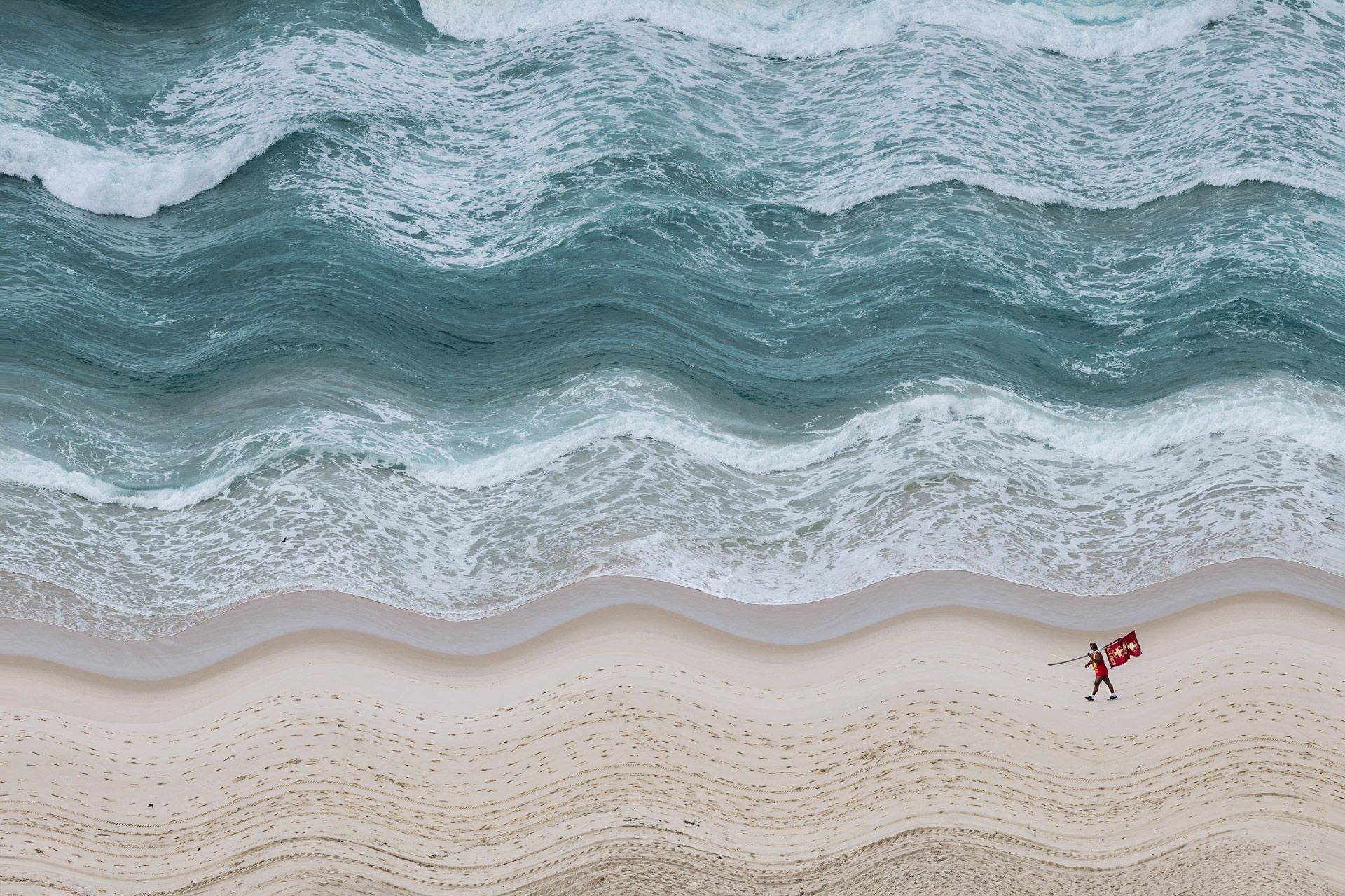 Vista aérea de ondas turquesas quebrando em uma praia de areia clara com uma pequena figura vermelha perto da costa.