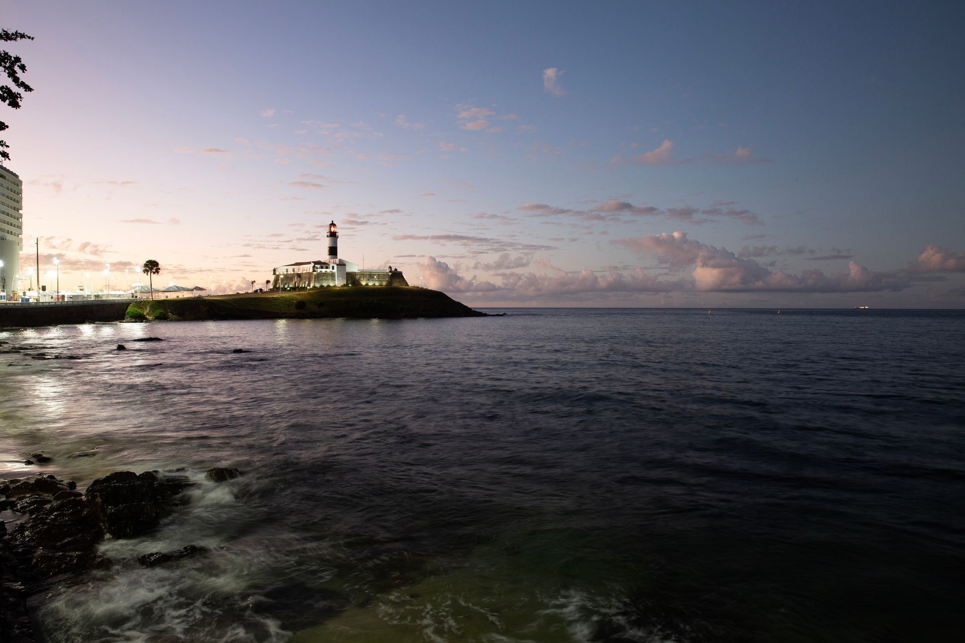 Pôr do sol sobre um oceano calmo ao lado de uma costa rochosa com um farol ao longe.