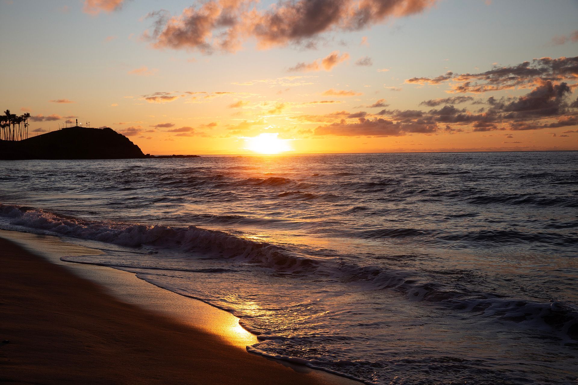 Pôr do sol sobre o oceano com ondas, um céu alaranjado brilhante e uma costa escura em silhueta.