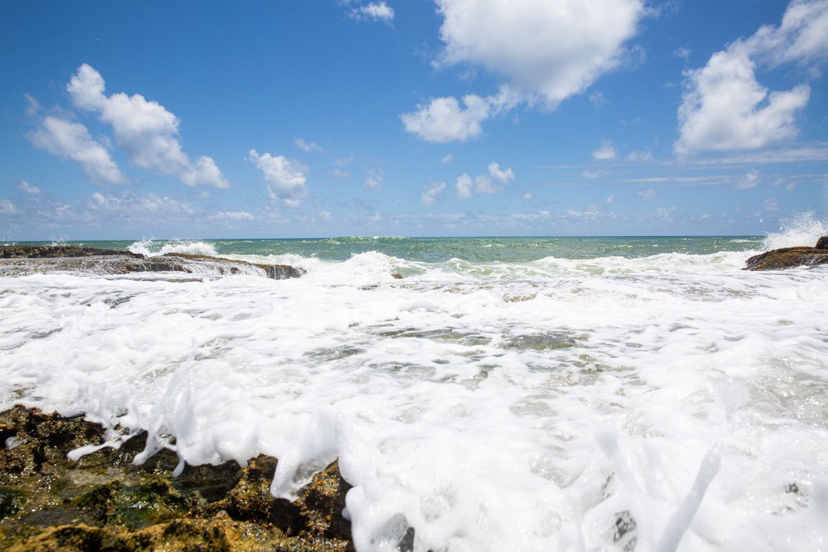 Ondas brancas e espumosas quebrando contra a costa rochosa sob um céu azul com nuvens dispersas.