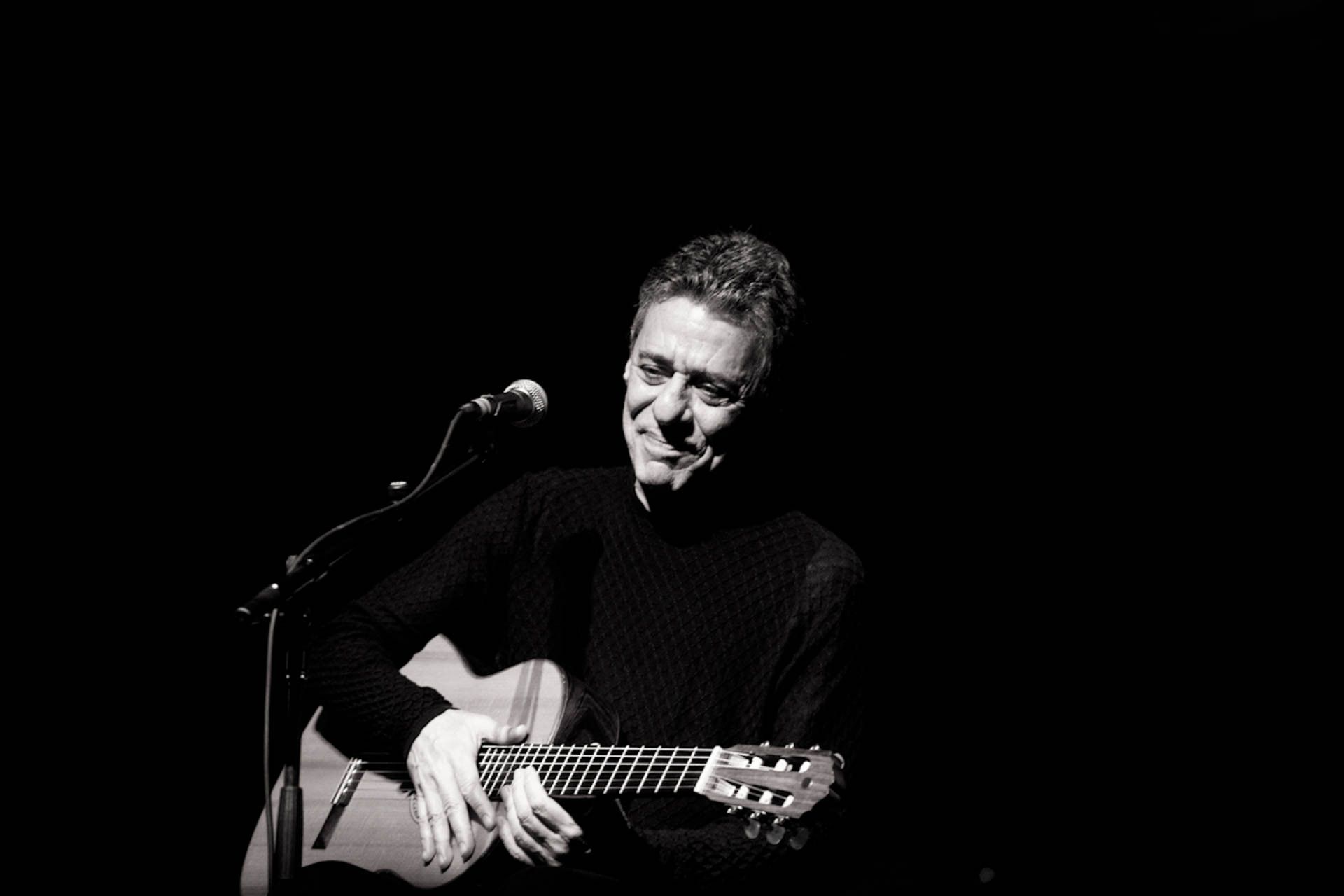 Músico em um palco escuro, sorrindo enquanto segura um violão ao lado de um microfone.