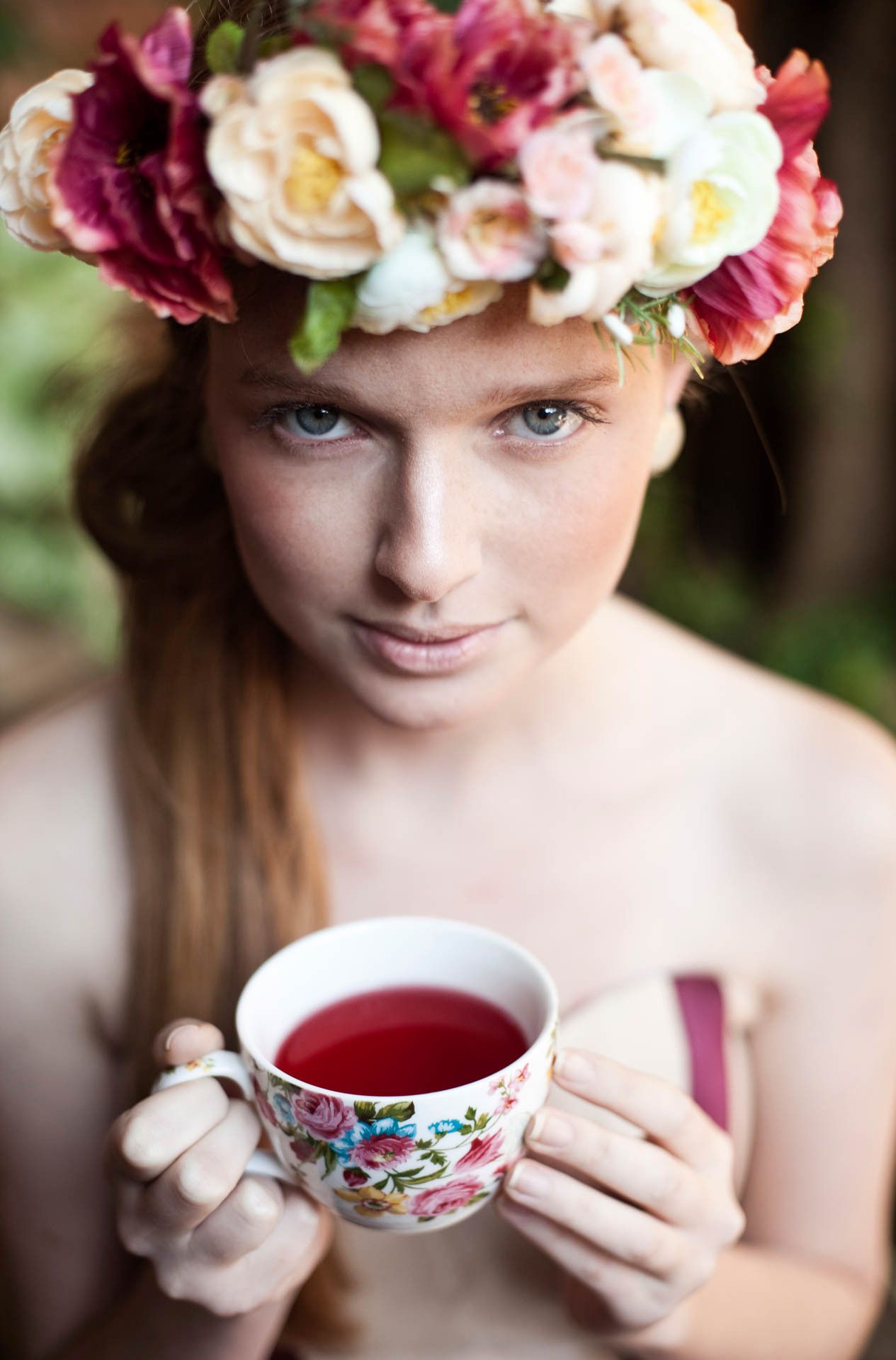 Mulher usando uma coroa de flores segura uma xícara de chá vermelha decorada com flores, olhando para a câmera.