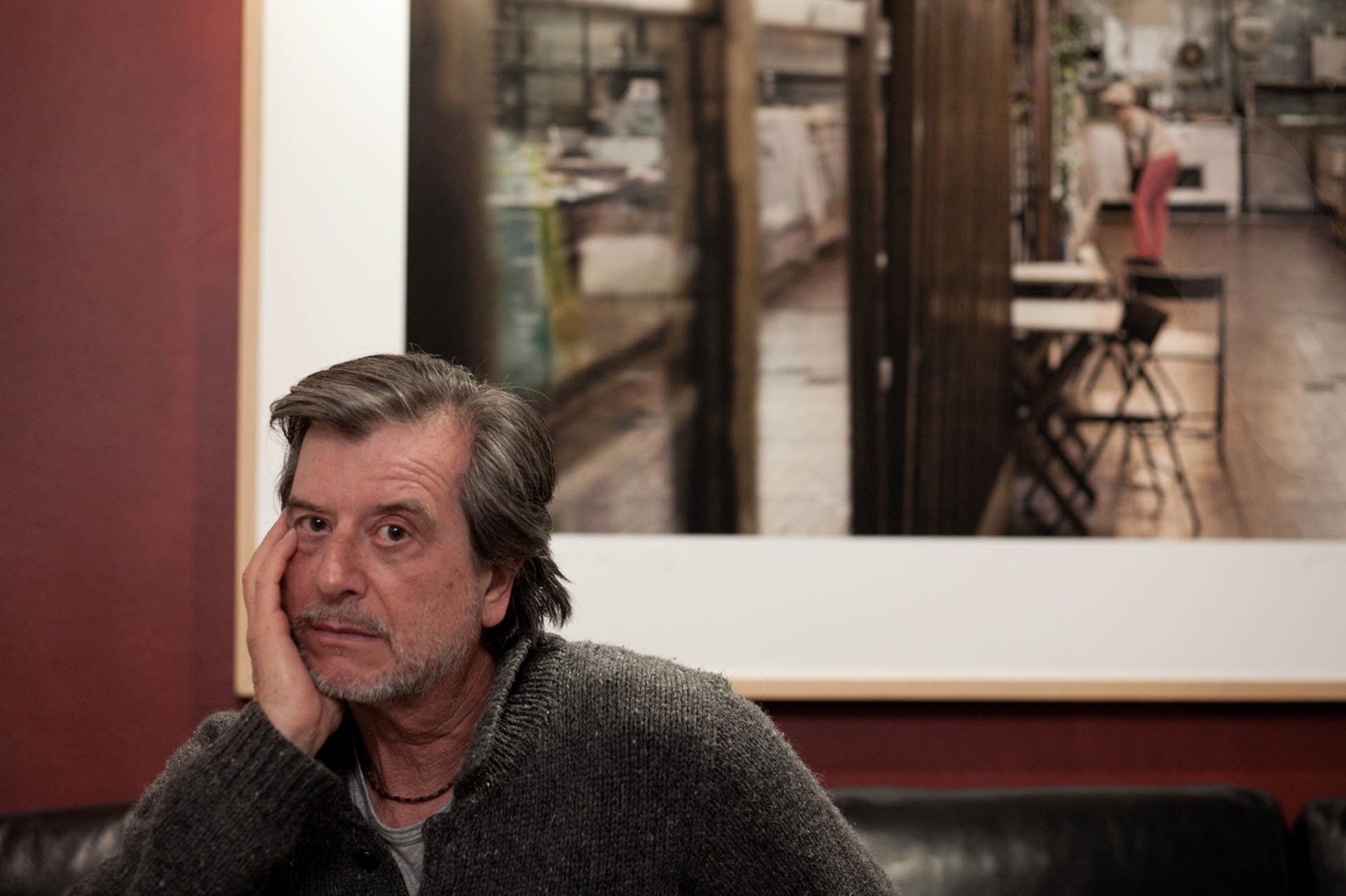 Homem sentado em um café, vestindo um suéter escuro, com uma foto emoldurada no corredor atrás dele.