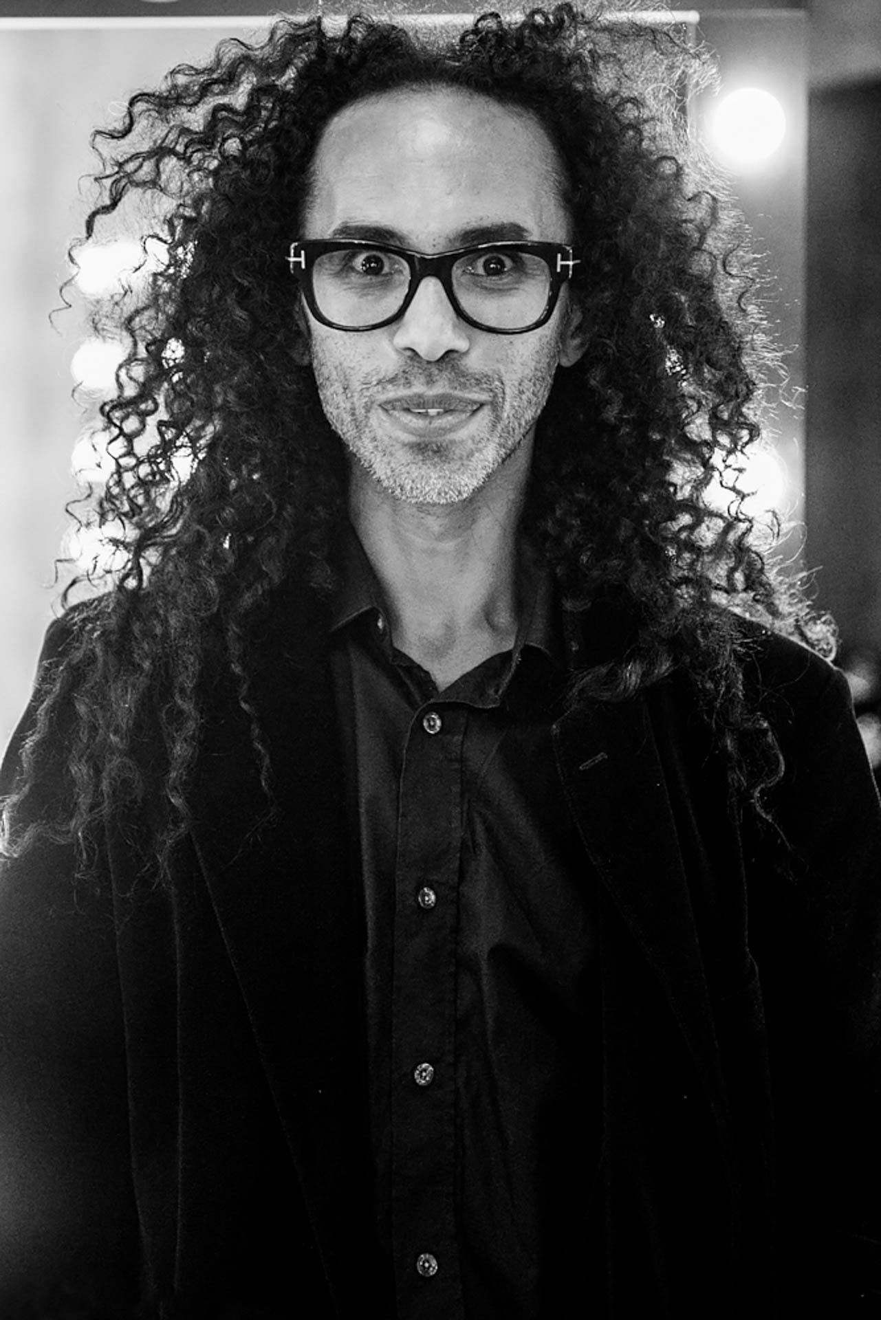 Retrato em preto e branco de uma pessoa com cabelo encaracolado e óculos, vestindo uma camisa escura, olhando para a câmera.