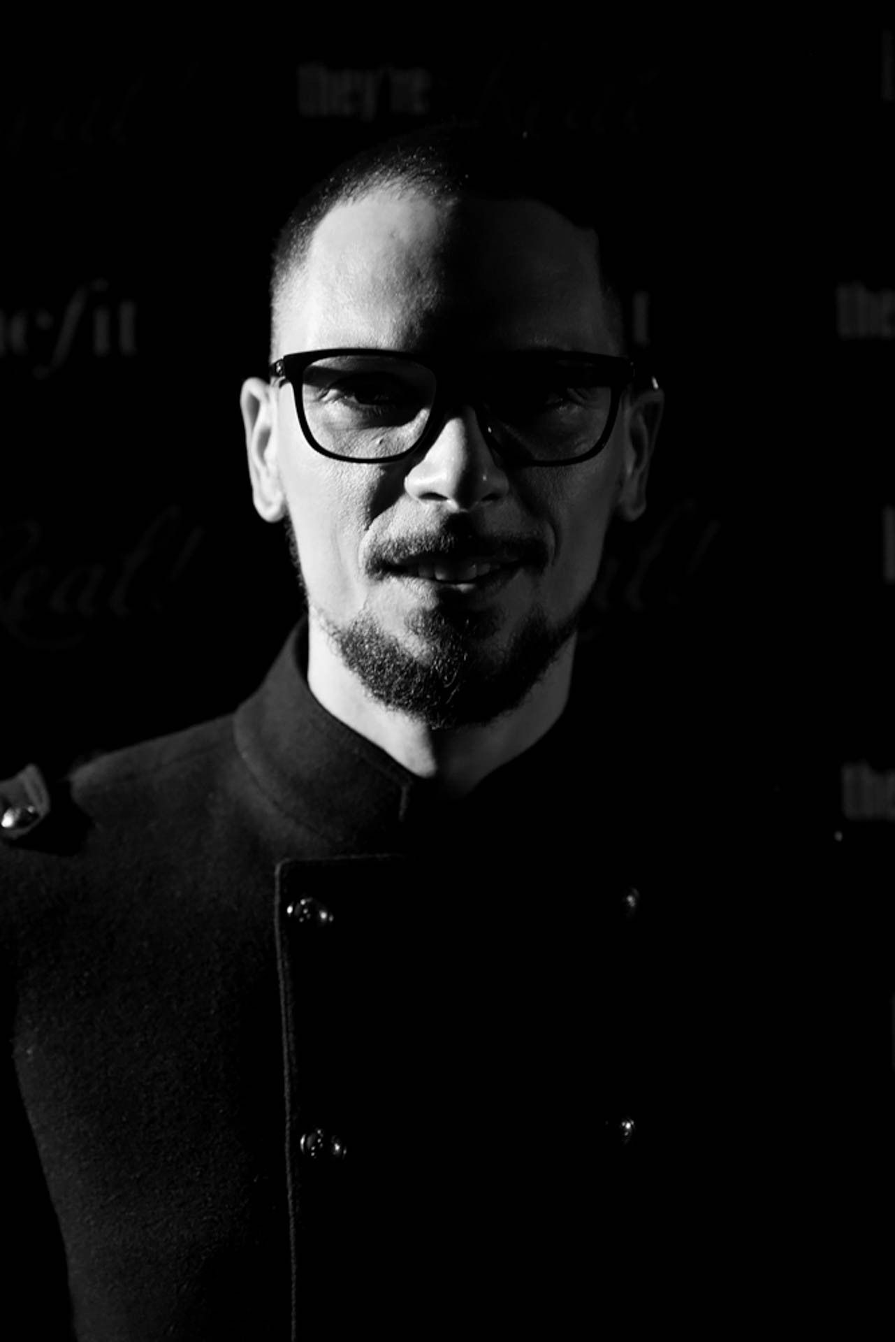 Retrato de um homem de óculos e roupas escuras contra um fundo preto, dramaticamente iluminado por um dos lados.