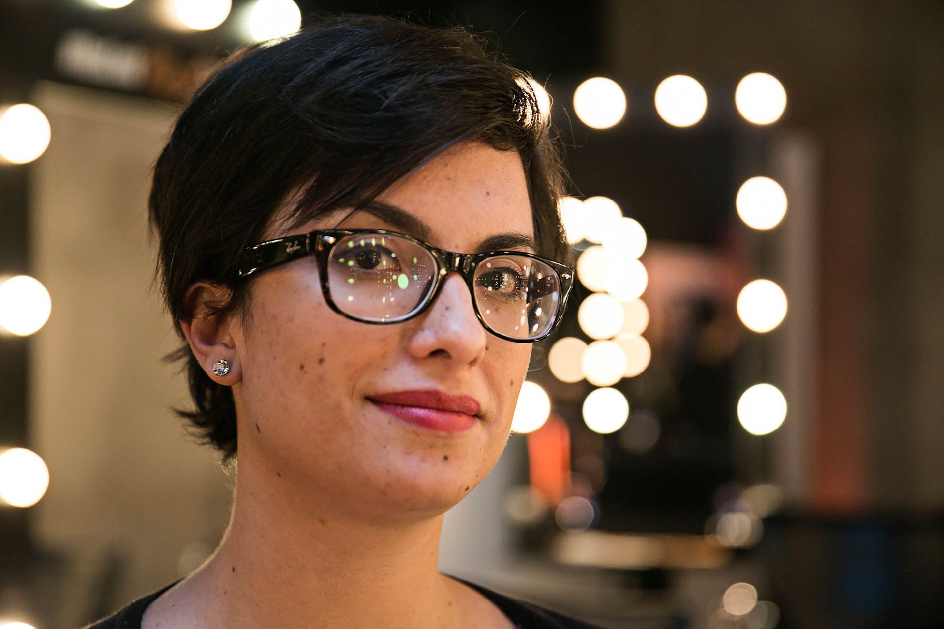 Retrato de uma pessoa com óculos e cabelo curto e escuro, suavemente iluminada contra luzes redondas desfocadas ao fundo.