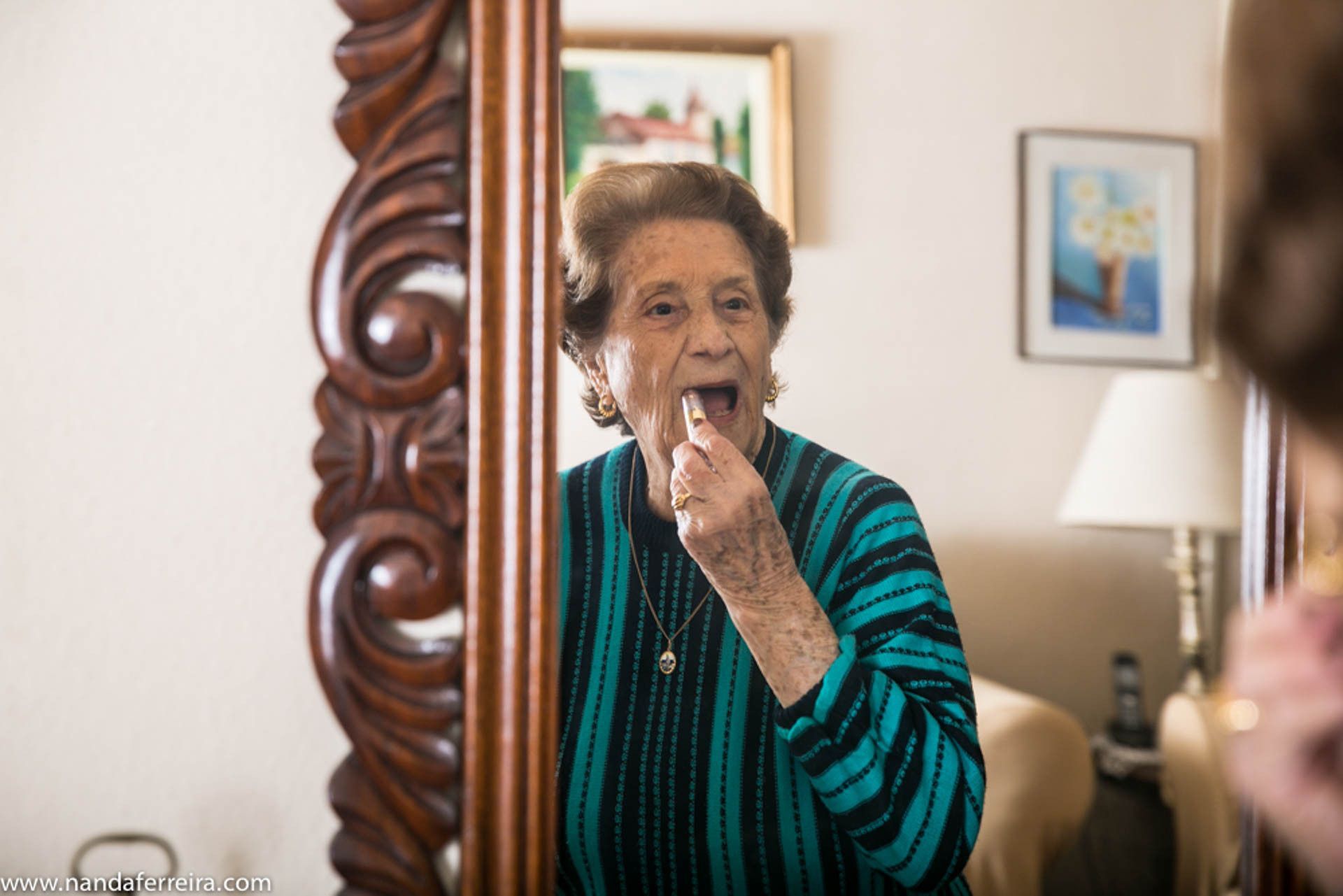 Mulher aplicando batom em frente ao espelho, vestindo um cardigã listrado em tons de azul-petróleo e preto.