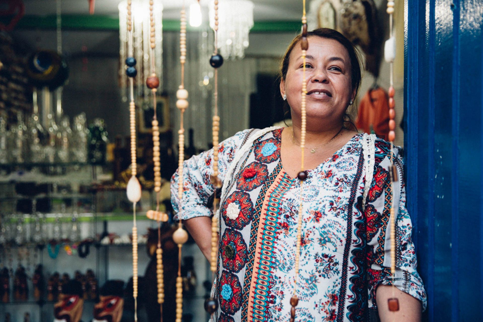 Mulher em pé na porta de uma loja com joias penduradas e roupas estampadas coloridas.