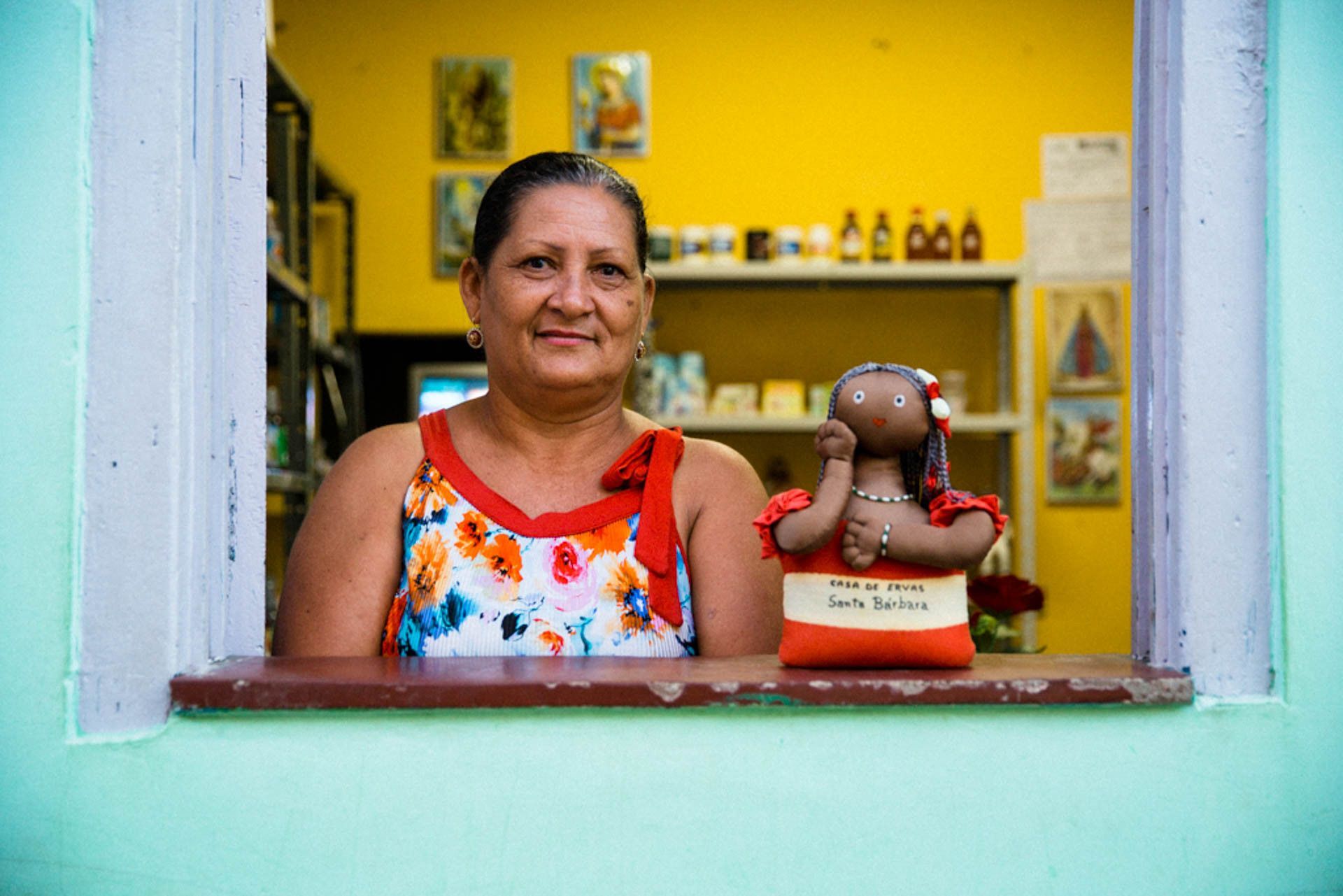 Mulher junto à janela de um quarto amarelo brilhante, em pé ao lado de uma pequena estatueta no parapeito.