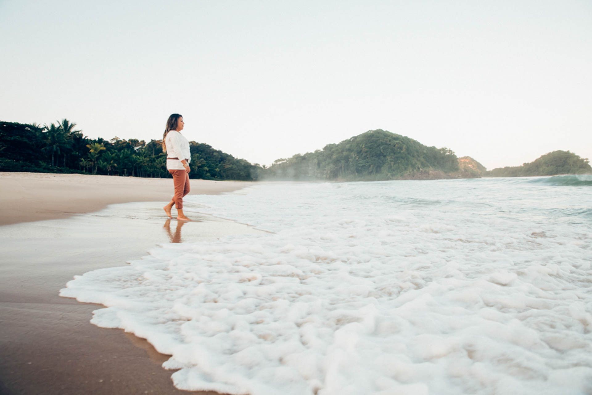 Pessoa caminhando ao longo de uma praia de areia ao lado de ondas espumantes, com colinas verdes ao fundo.