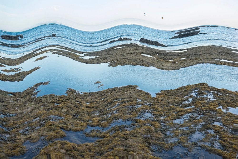 Reflexo ondulado de uma montanha nevada sobre águas azuis calmas, com costa rochosa em primeiro plano.