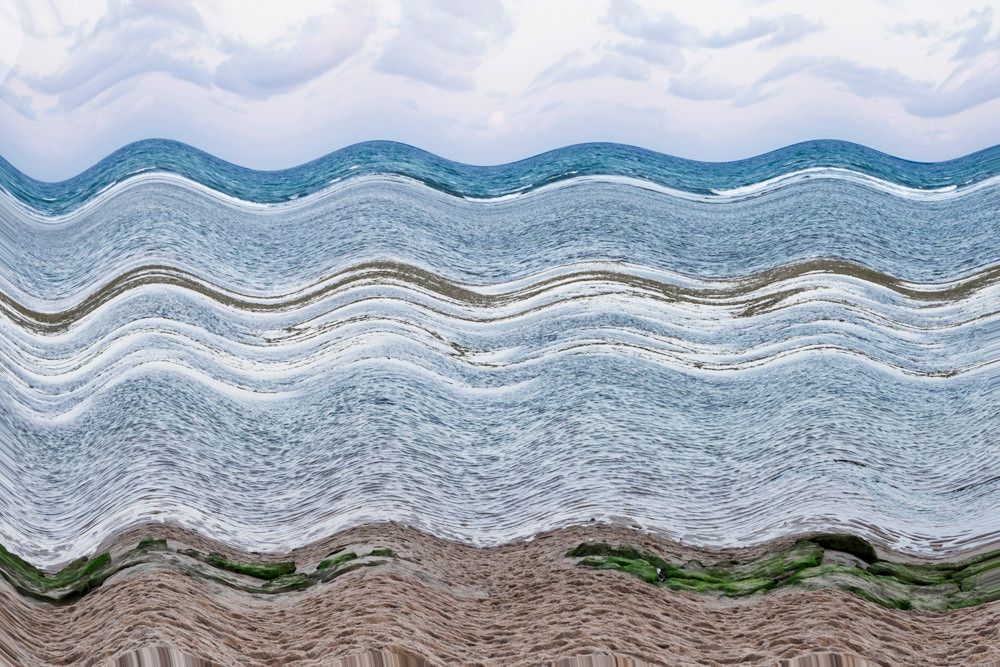 Paisagem litorânea ondulada com água azul, ondas brancas e praia de areia sob um céu nublado.