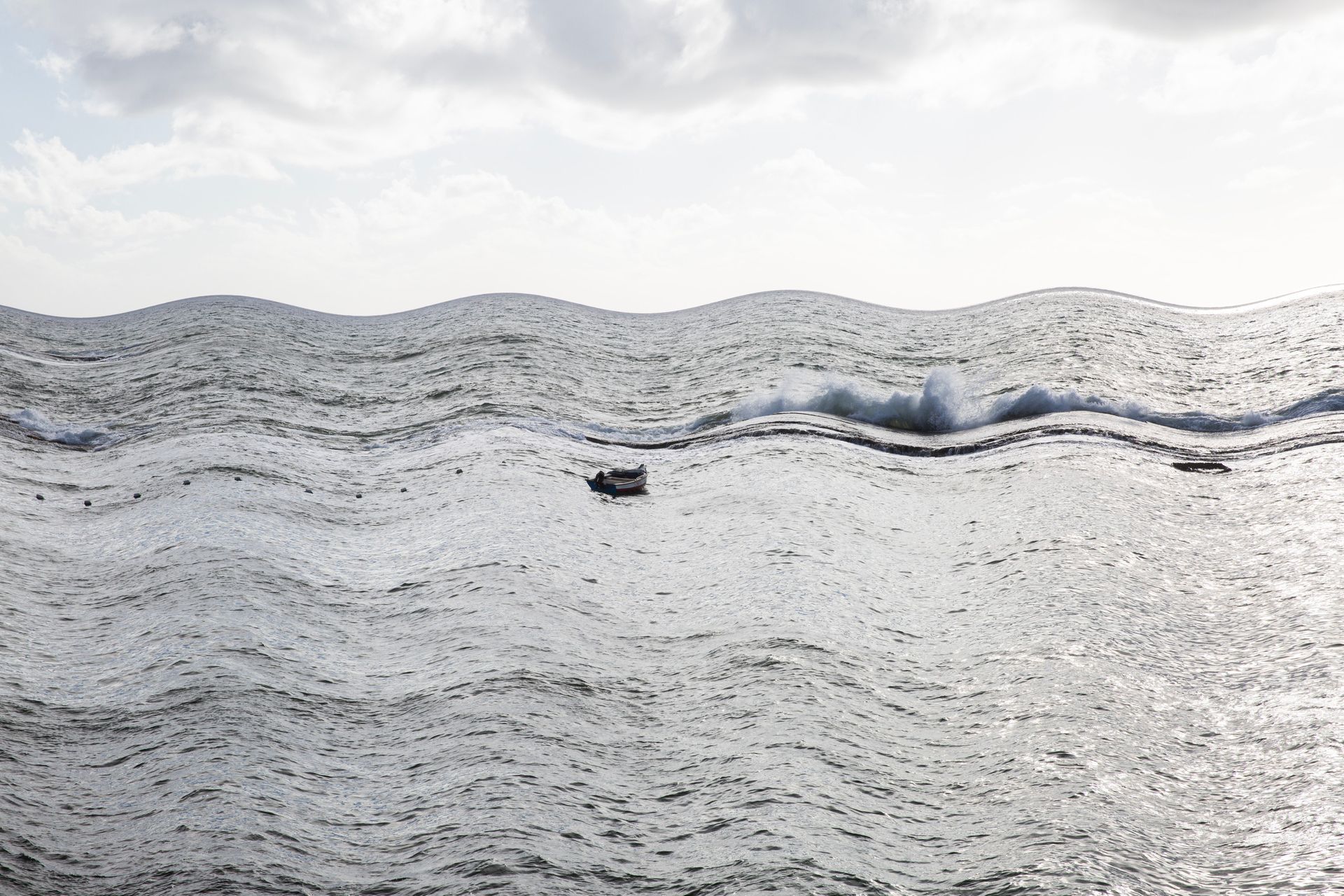 Pequeno barco em águas cinzentas e agitadas, com uma onda quebrando ao longe sob um céu nublado.