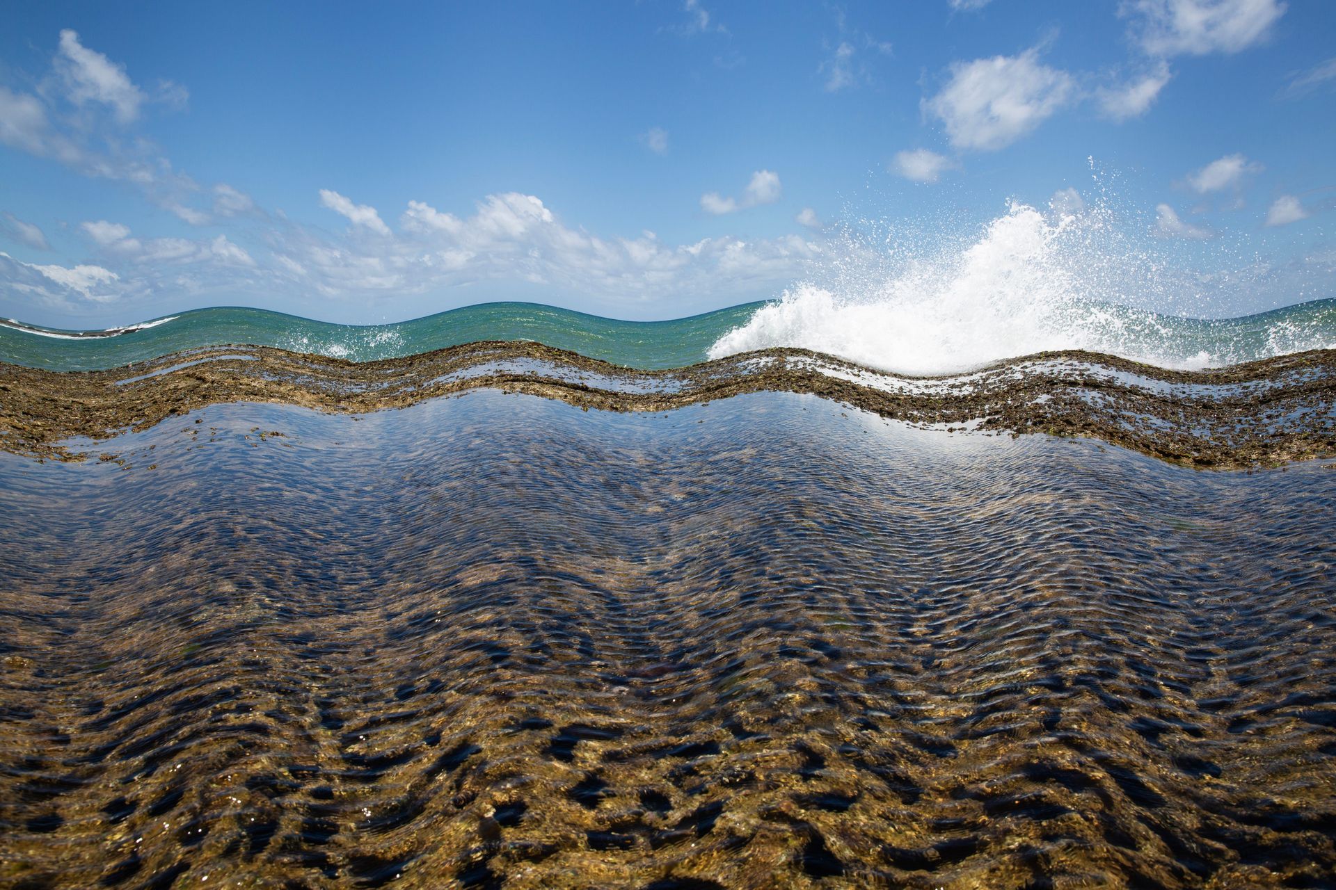 Águas cristalinas e rasas do mar, com seixos ondulados, ondas e um céu azul com nuvens.
