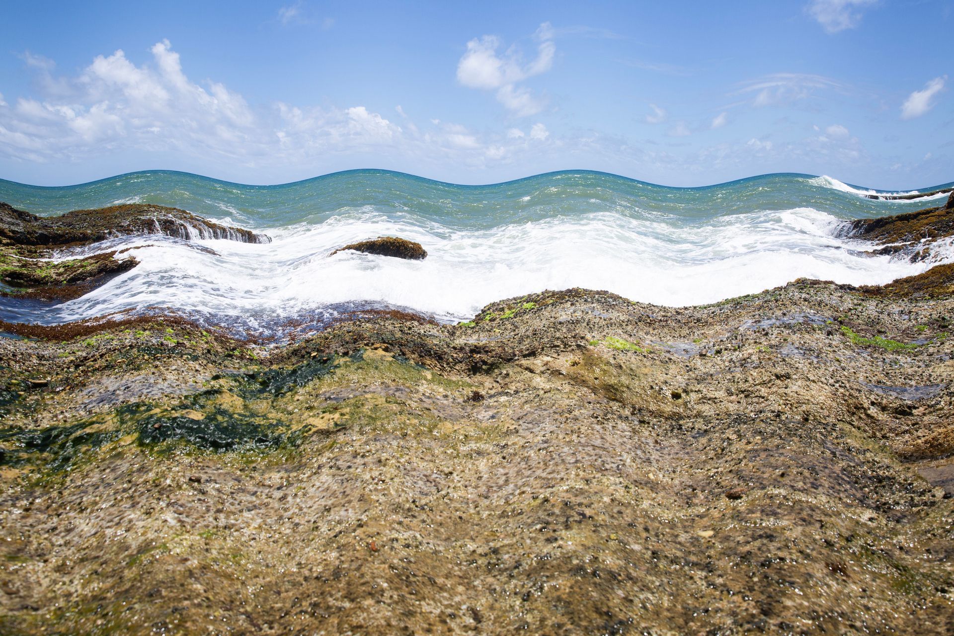 Ondas rasas do oceano quebrando sobre uma costa rochosa e arenosa sob um céu azul.