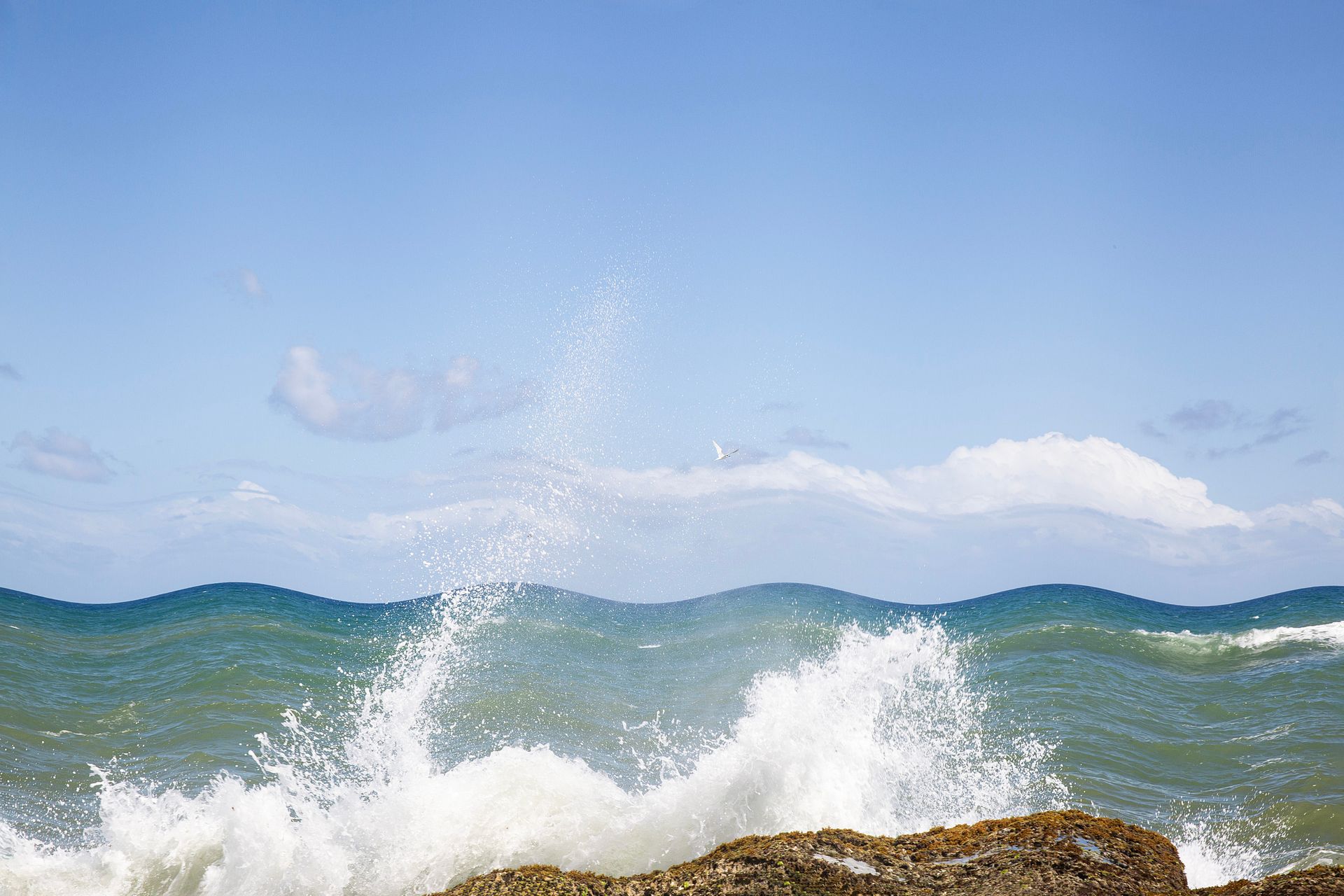 Ondas quebrando em uma costa rochosa sob um céu azul claro com nuvens distantes.