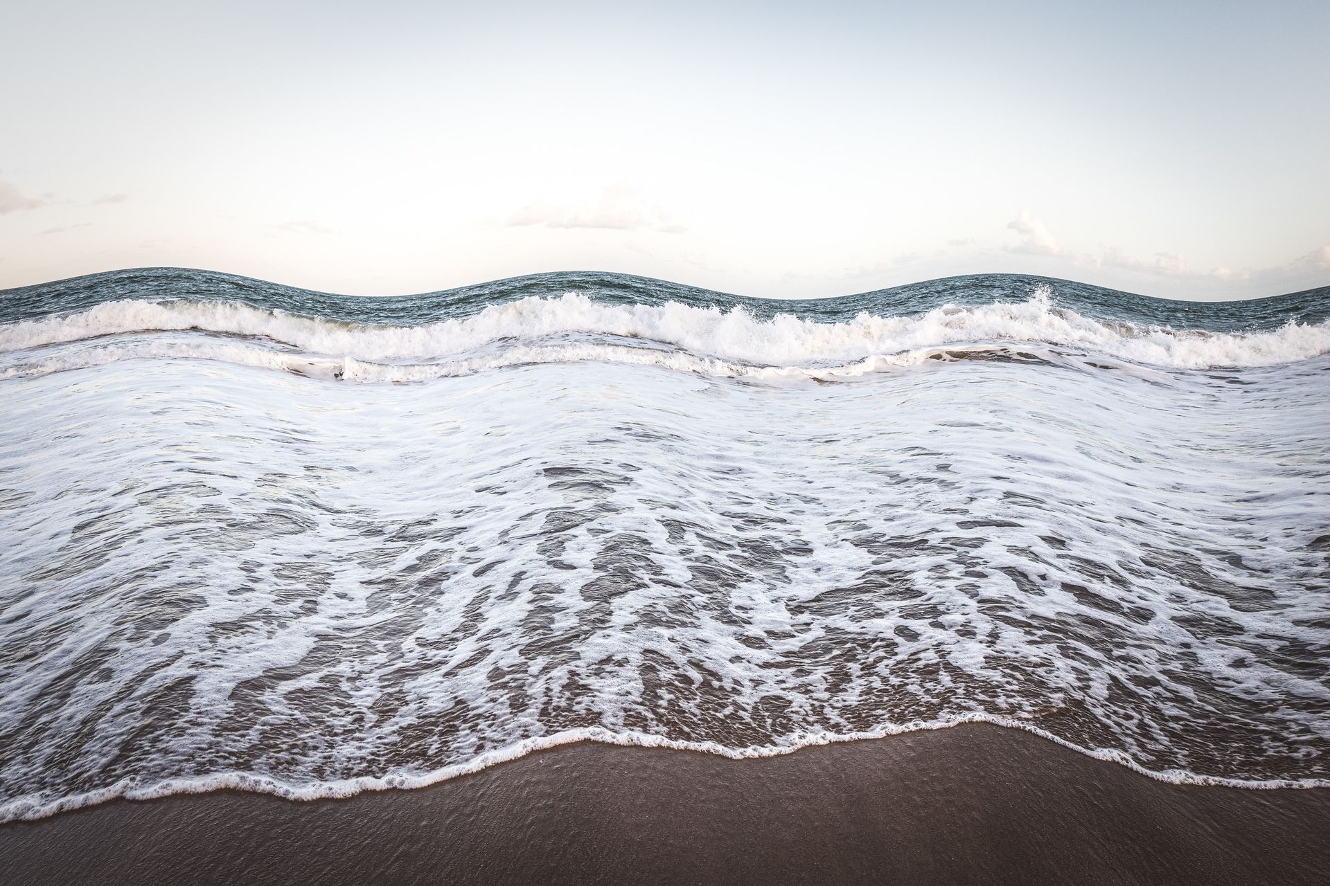 Ondas suaves do oceano banhando uma praia de areia sob um céu pálido.