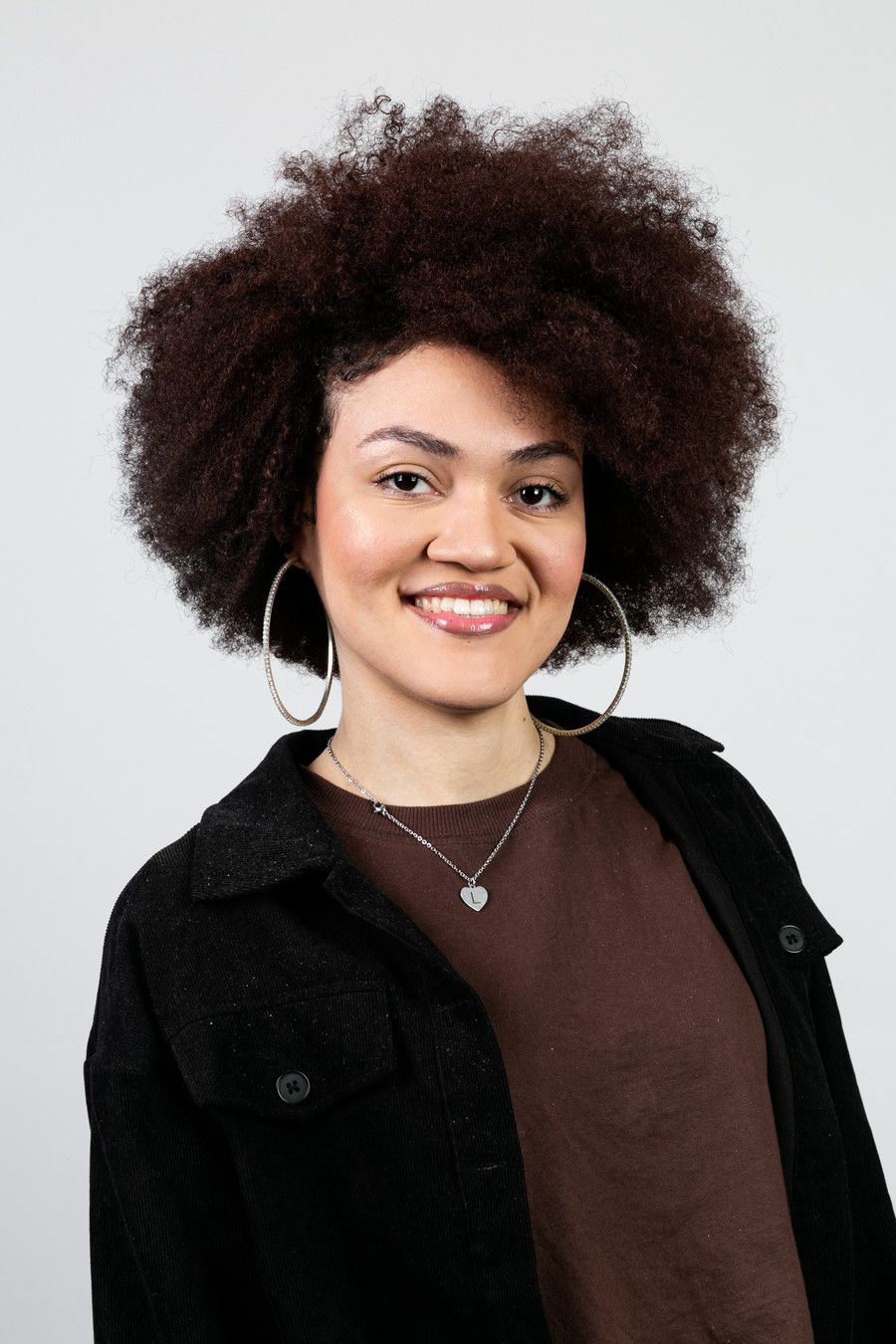 Pessoa sorridente com cabelo encaracolado, vestindo uma jaqueta preta sobre uma blusa marrom, em um fundo claro.