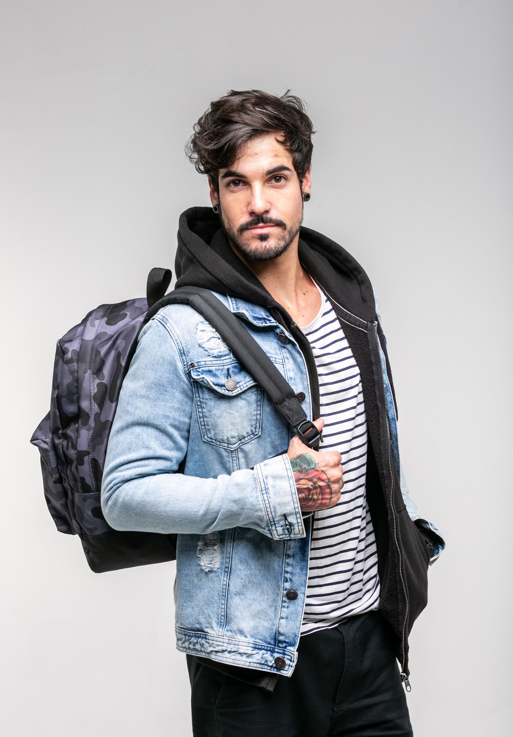 Homem vestindo jaqueta jeans e mochila, em pé contra um fundo cinza claro.