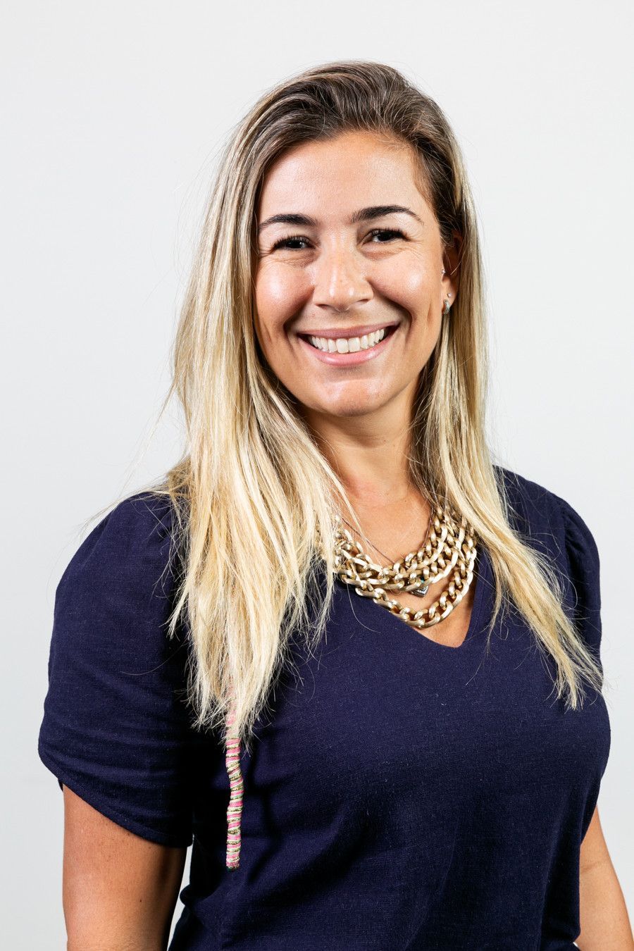 Mulher sorridente com blusa azul-marinho e colar dourado contra um fundo branco.