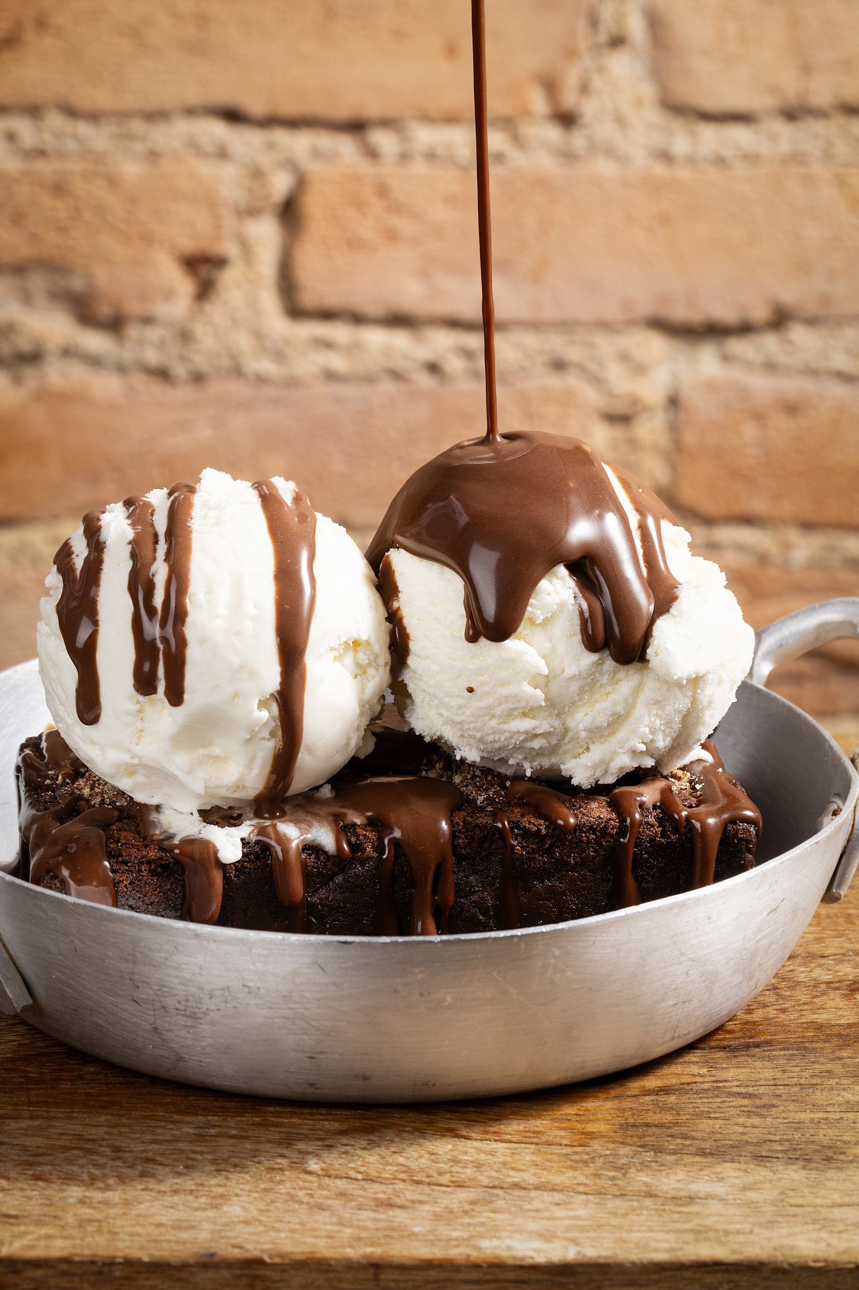 Duas bolas de sorvete de baunilha sobre bolo de chocolate, regadas com calda de chocolate em uma travessa de metal.