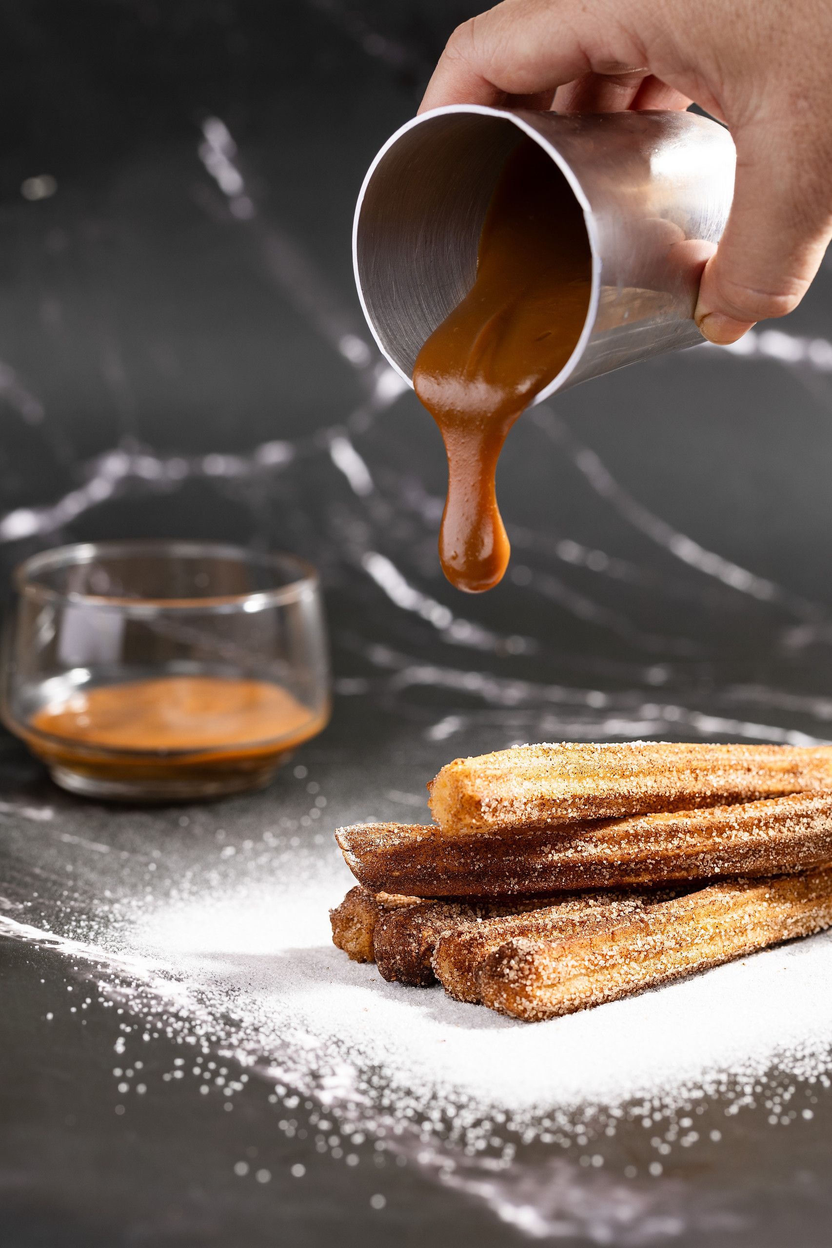 Derramar calda de caramelo à mão sobre uma pilha de churros em uma mesa escura com açúcar de confeiteiro.
