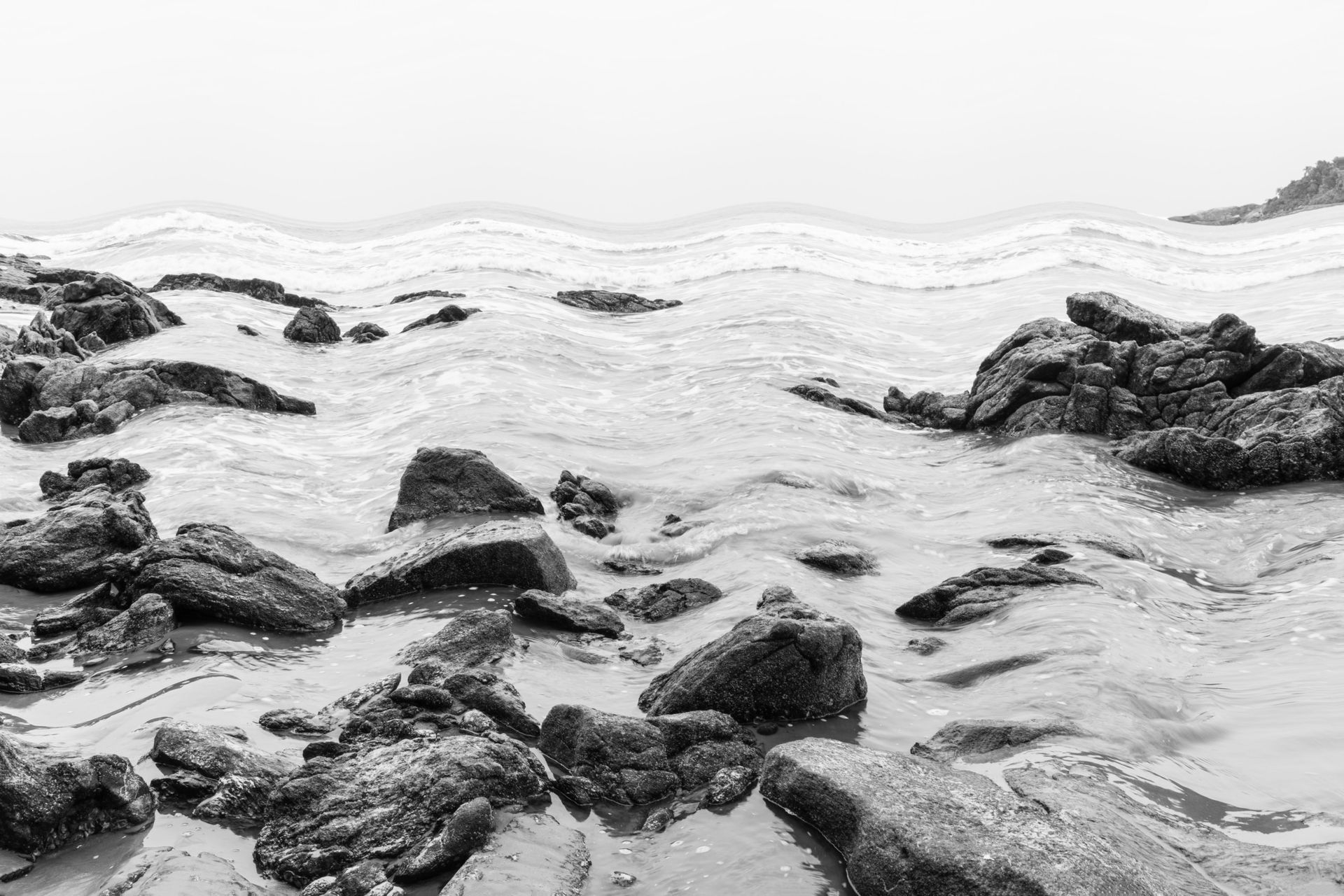 Costa rochosa em preto e branco com ondas quebrando sobre pedras molhadas sob um céu enevoado.