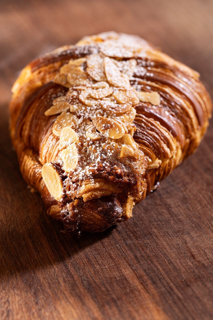 Um croissant de amêndoa dourado sobre uma superfície de madeira, coberto com amêndoas laminadas e açúcar de confeiteiro.