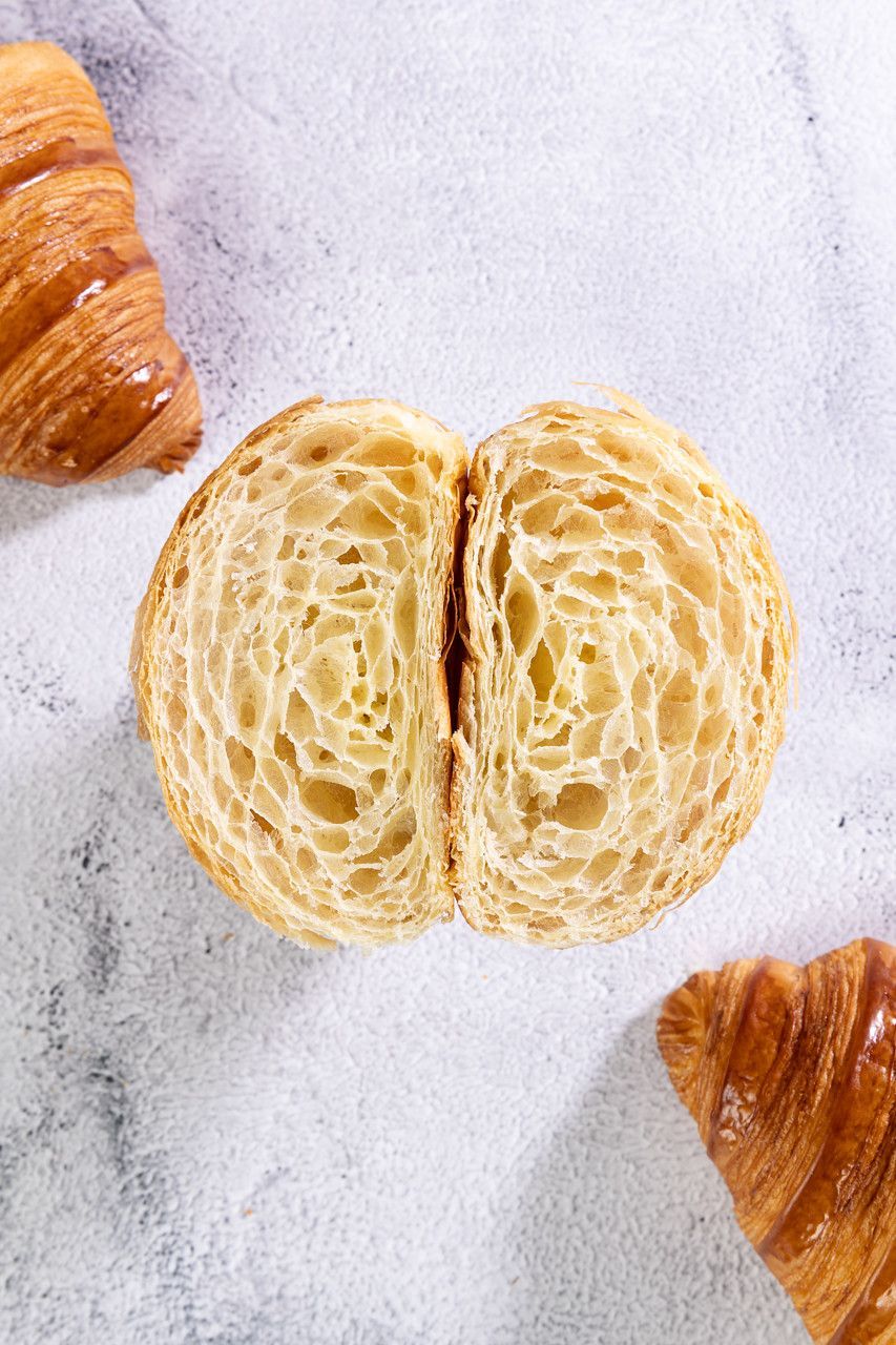 Croissant fatiado com massa folhada sobre uma superfície levemente texturizada, com croissants inteiros parcialmente visíveis nas proximidades.