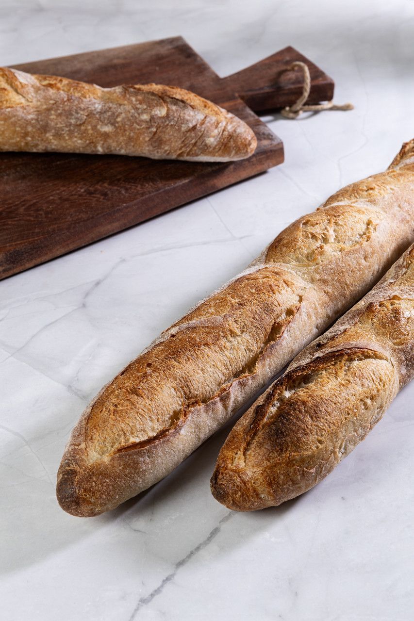 Duas baguetes crocantes sobre uma superfície de mármore ao lado de uma tábua de corte de madeira, com uma faca para baguete ao fundo.
