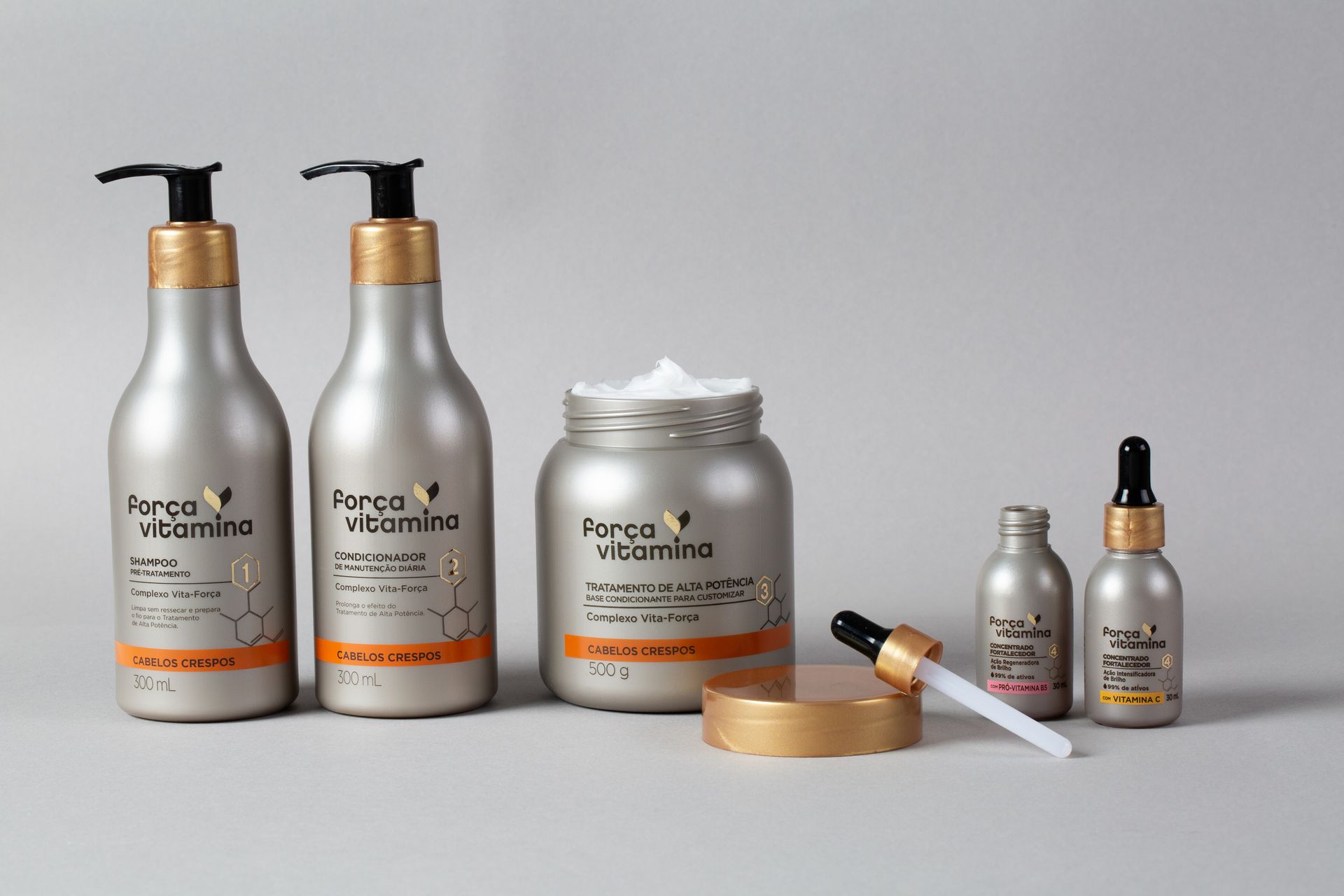 Frascos e pote de produtos para cuidados com a pele prateados com detalhes dourados, além de frascos conta-gotas e espátula sobre um fundo cinza.