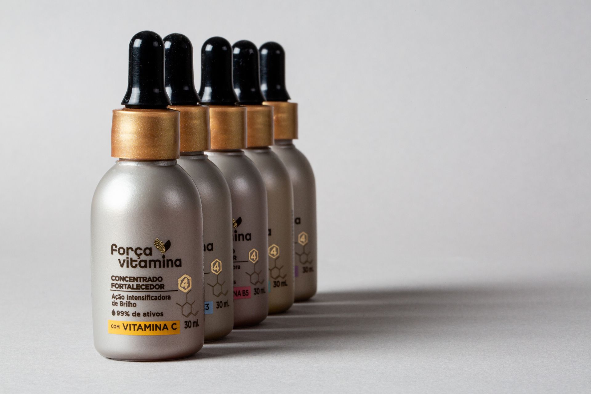 Três frascos conta-gotas de vitaminas foscos com tampas pretas e anéis dourados sobre um fundo cinza.