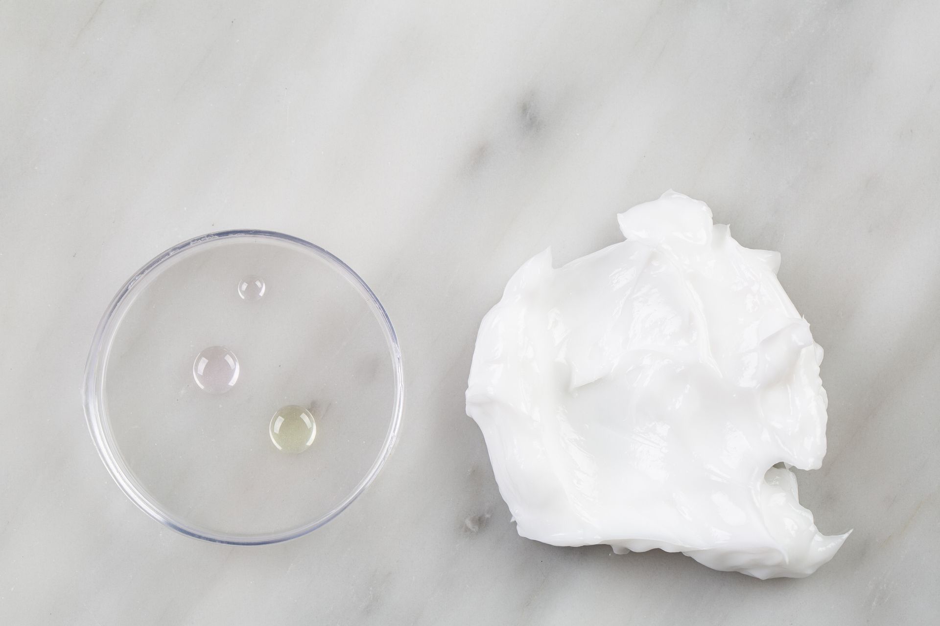 Três gotas transparentes em uma placa de Petri ao lado de um papel branco sobre uma superfície de mármore.