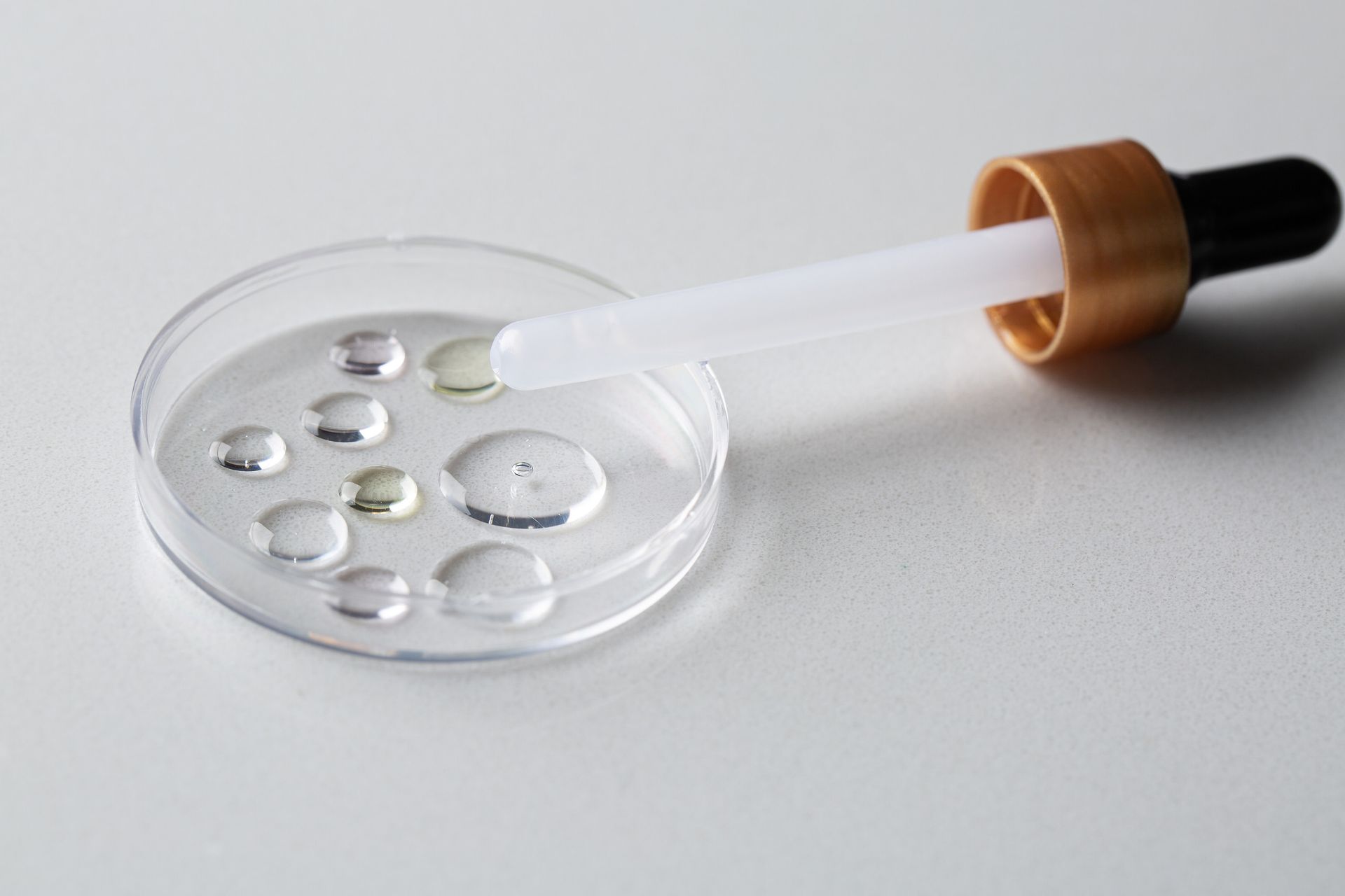 Conta-gotas de vidro dispensando líquido transparente em uma placa de Petri com gotas de água.