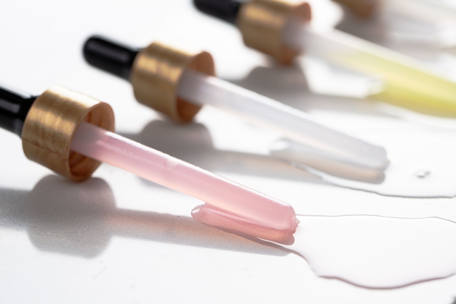 Imagem em close-up de pipetas de vidro com líquido rosa, branco e amarelo sobre uma superfície branca.