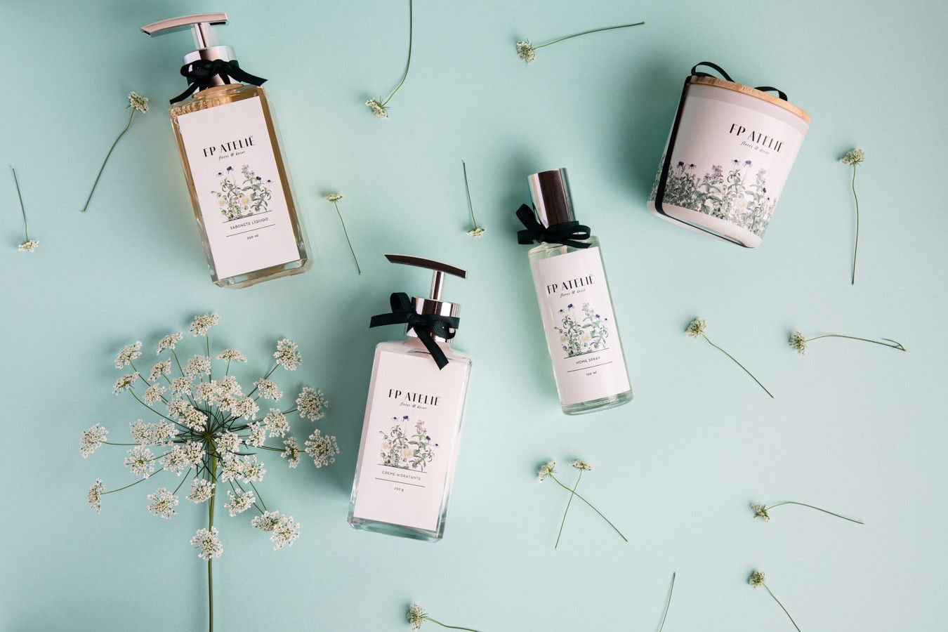 Três frascos brancos de produtos para cuidados com a pele com estampa floral sobre um fundo azul claro com flores secas e raminhos.