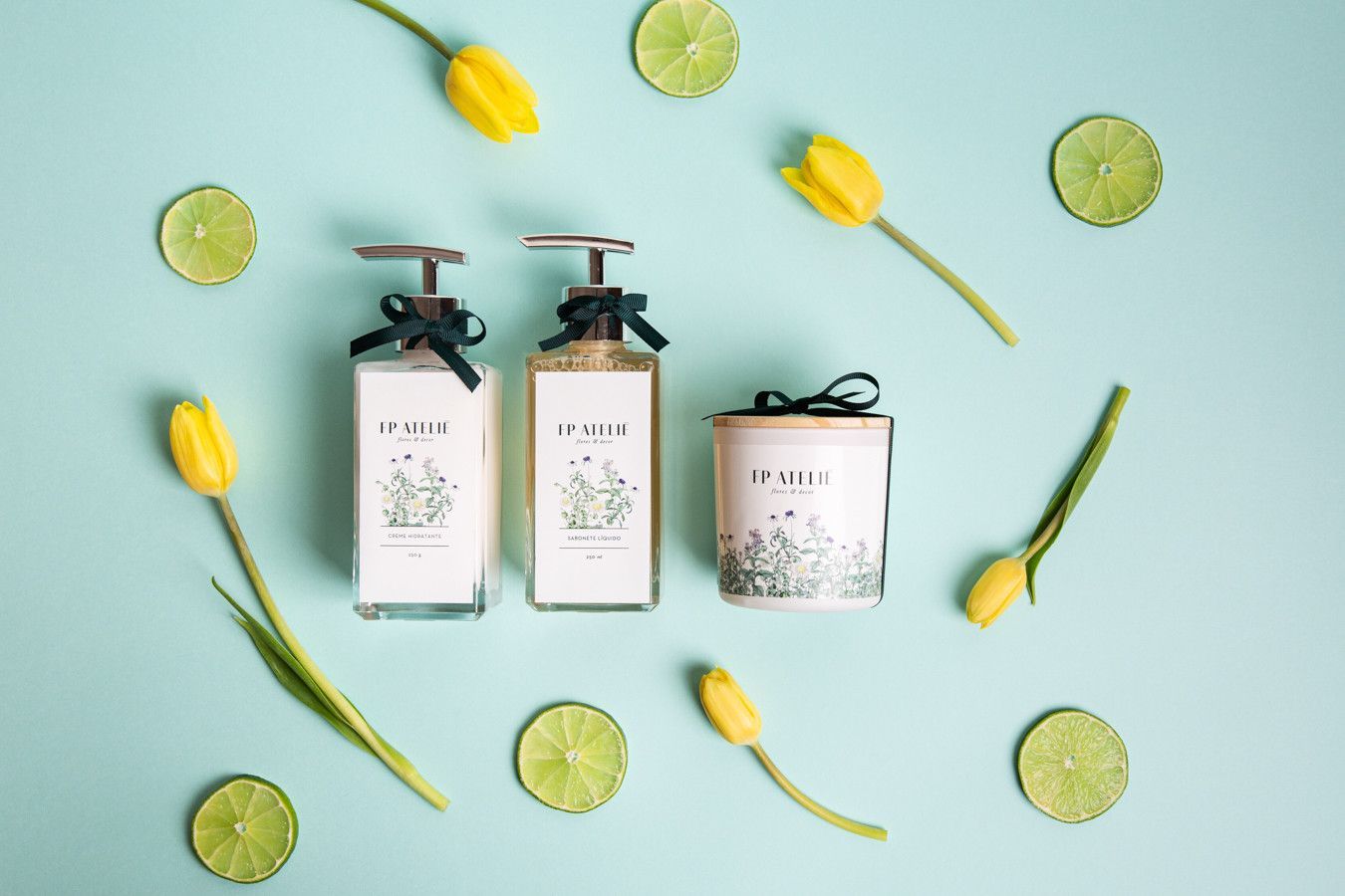 Três frascos de sabonete branco e uma vela sobre uma superfície azul-clara com limões e tulipas amarelas.