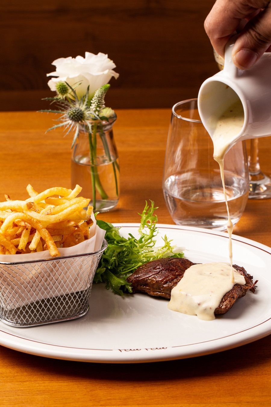 Bife com batatas fritas servido à mesa do restaurante com molho, acompanhado de uma taça de vinho e flores.
