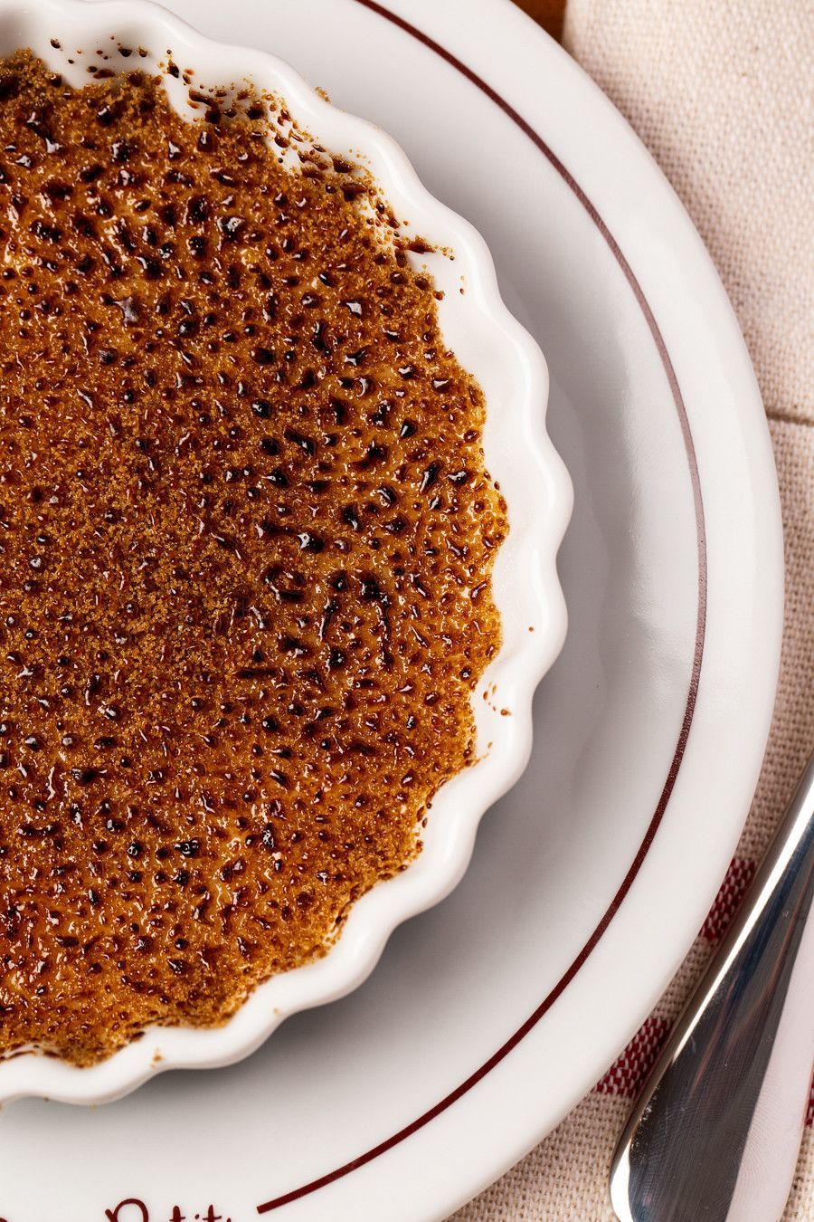 Crème brûlée branco com bordas onduladas e crosta caramelizada, servido em um prato, ao lado de uma colher sobre um guardanapo bege.