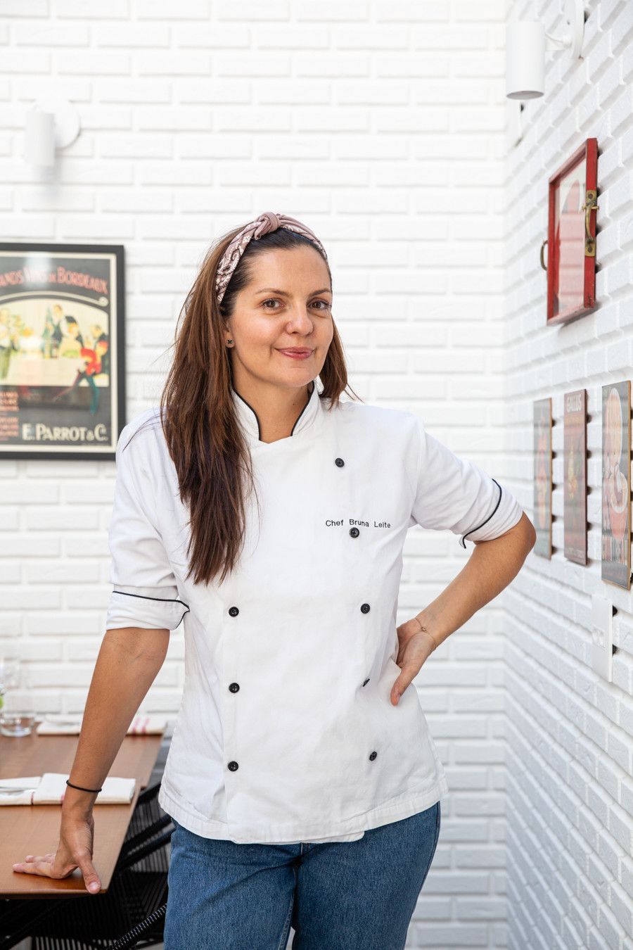 Mulher com um casaco de chef branco em pé na cozinha de um restaurante com paredes de tijolos bem iluminadas.